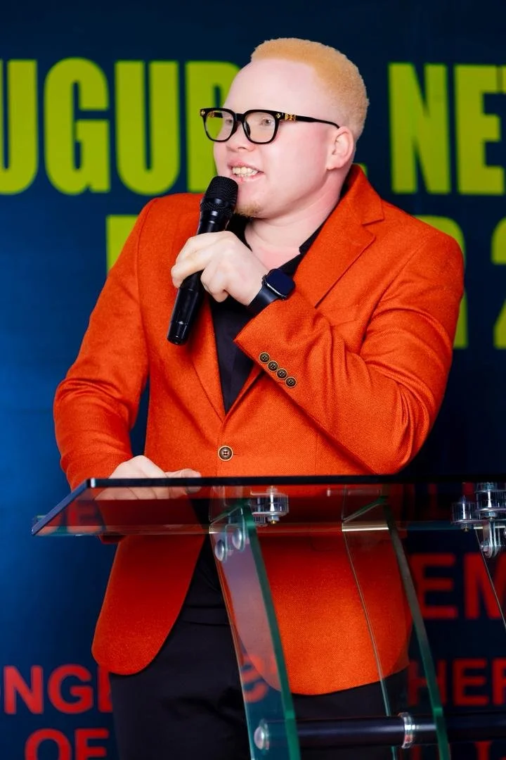 A person with short blonde hair, glasses, and a red blazer speaking into a microphone behind a clear podium.