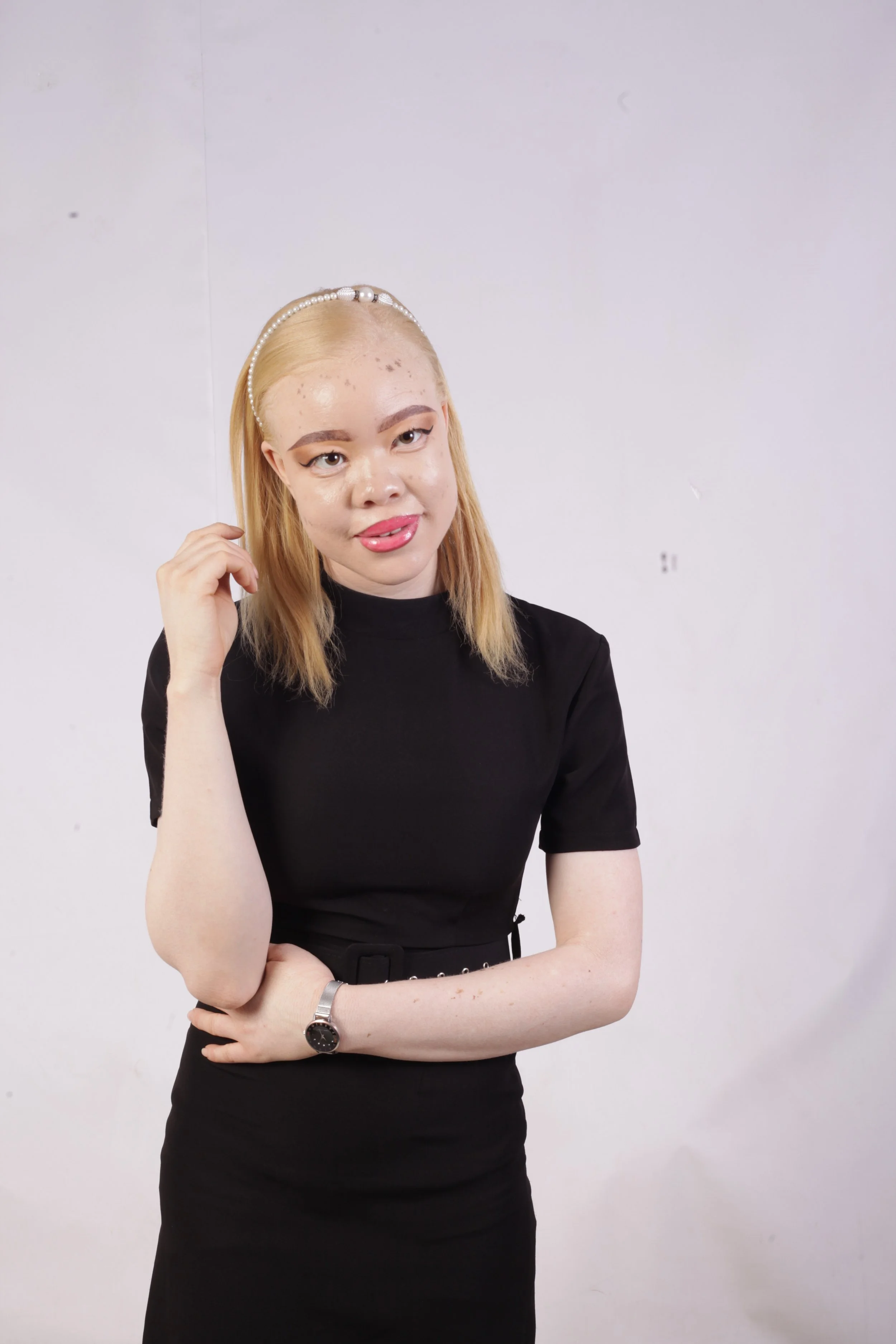 A young woman with blonde hair and freckles, wearing a black dress with a belt, a pearl headband, and a watch, standing against a plain white background.