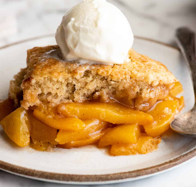 A slice of peach cobbler with a scoop of vanilla ice cream on top, served on a white plate with a spoon.