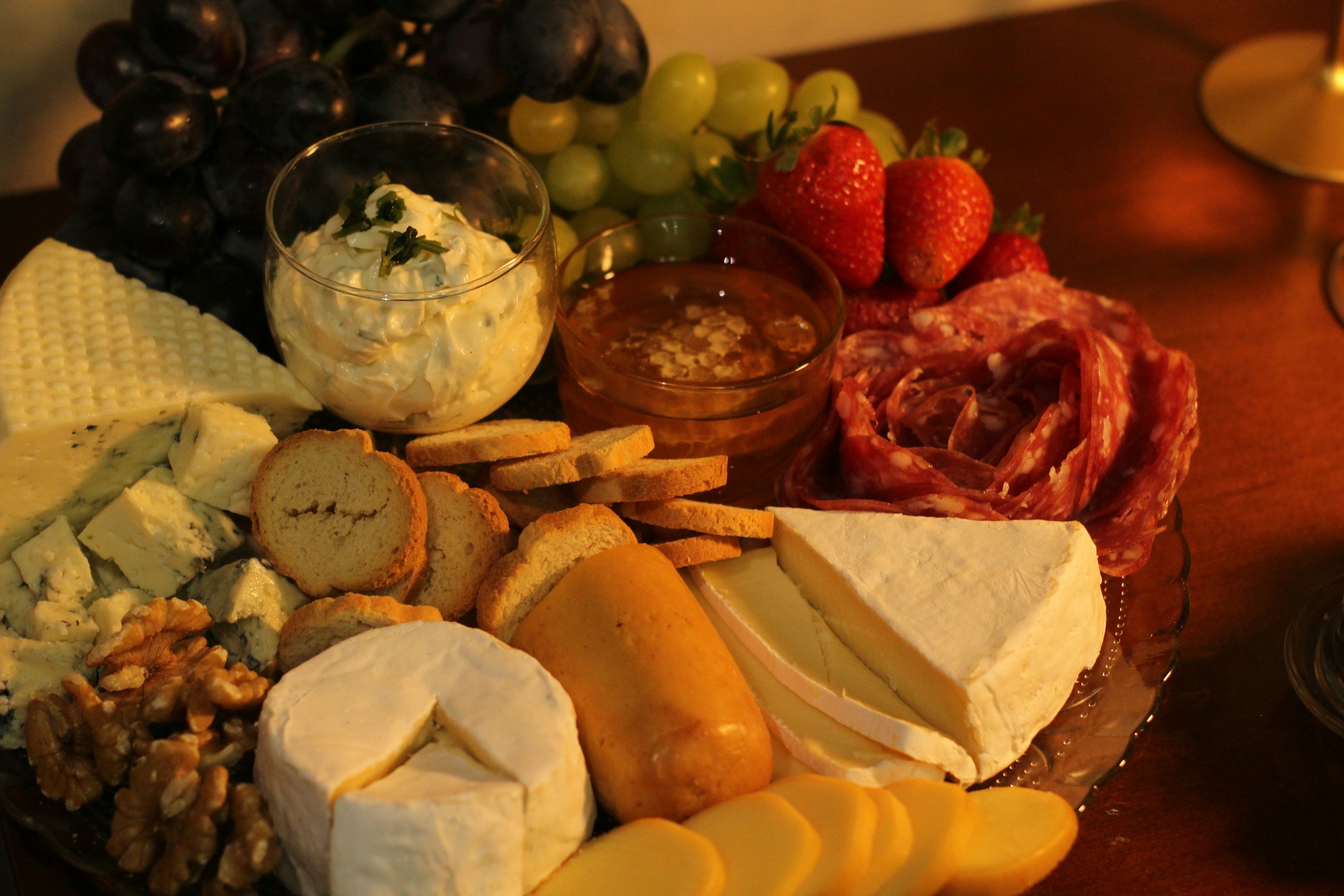 A cheese and charcuterie board featuring various cheeses, grapes, strawberries, salami, crackers, breadsticks, a dip, whipped butter, and walnuts on a wooden table.