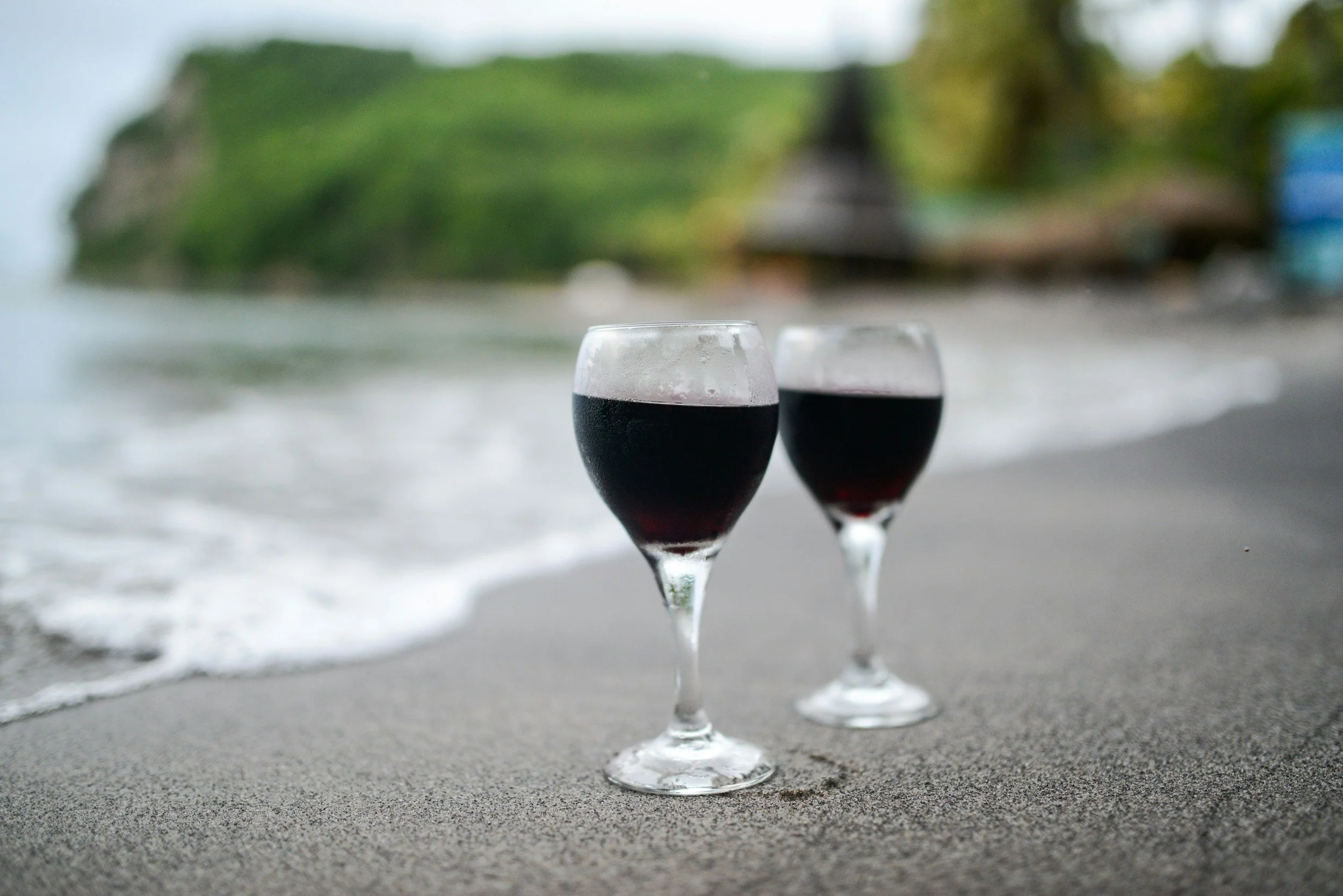 Two glasses of red wine on the sandy beach with ocean waves and green hills in the background.