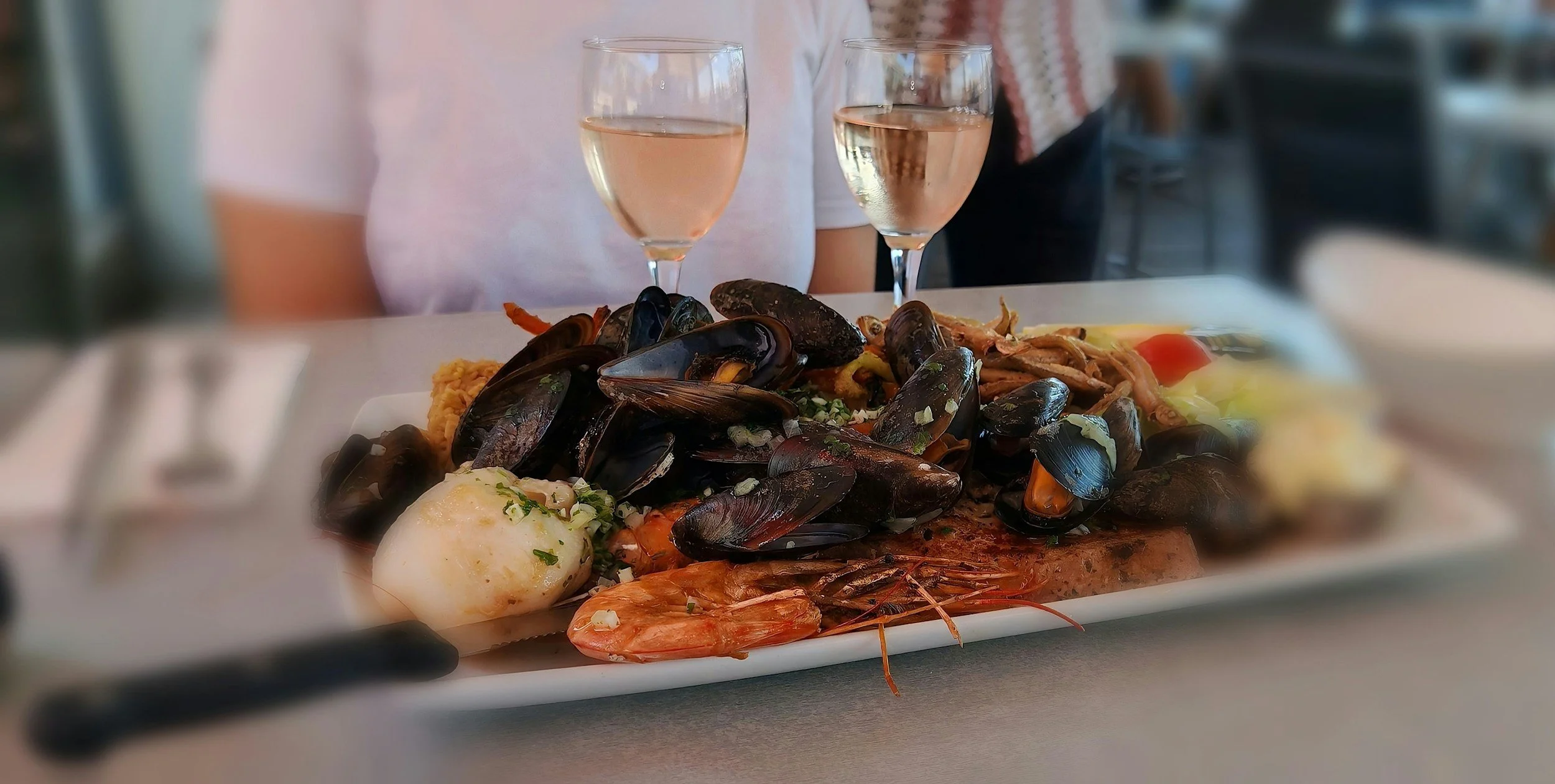 Seafood platter with mussels, shrimp, and other shellfish, served with a poached egg and garnished with herbs, accompanied by two glasses of white wine.