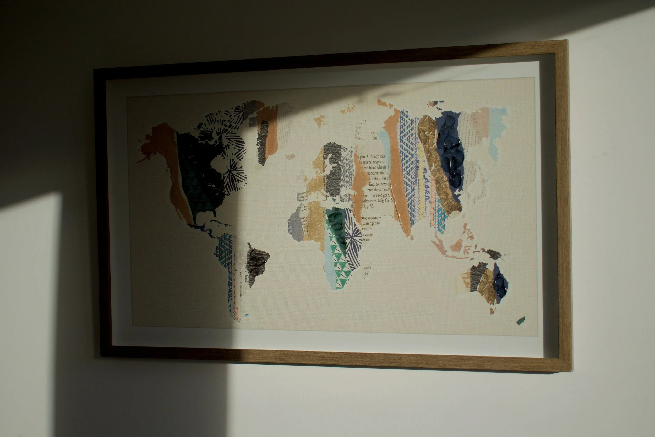 A framed world map collage made of torn paper and magazine clippings, hanging on a wall, partially illuminated by sunlight.