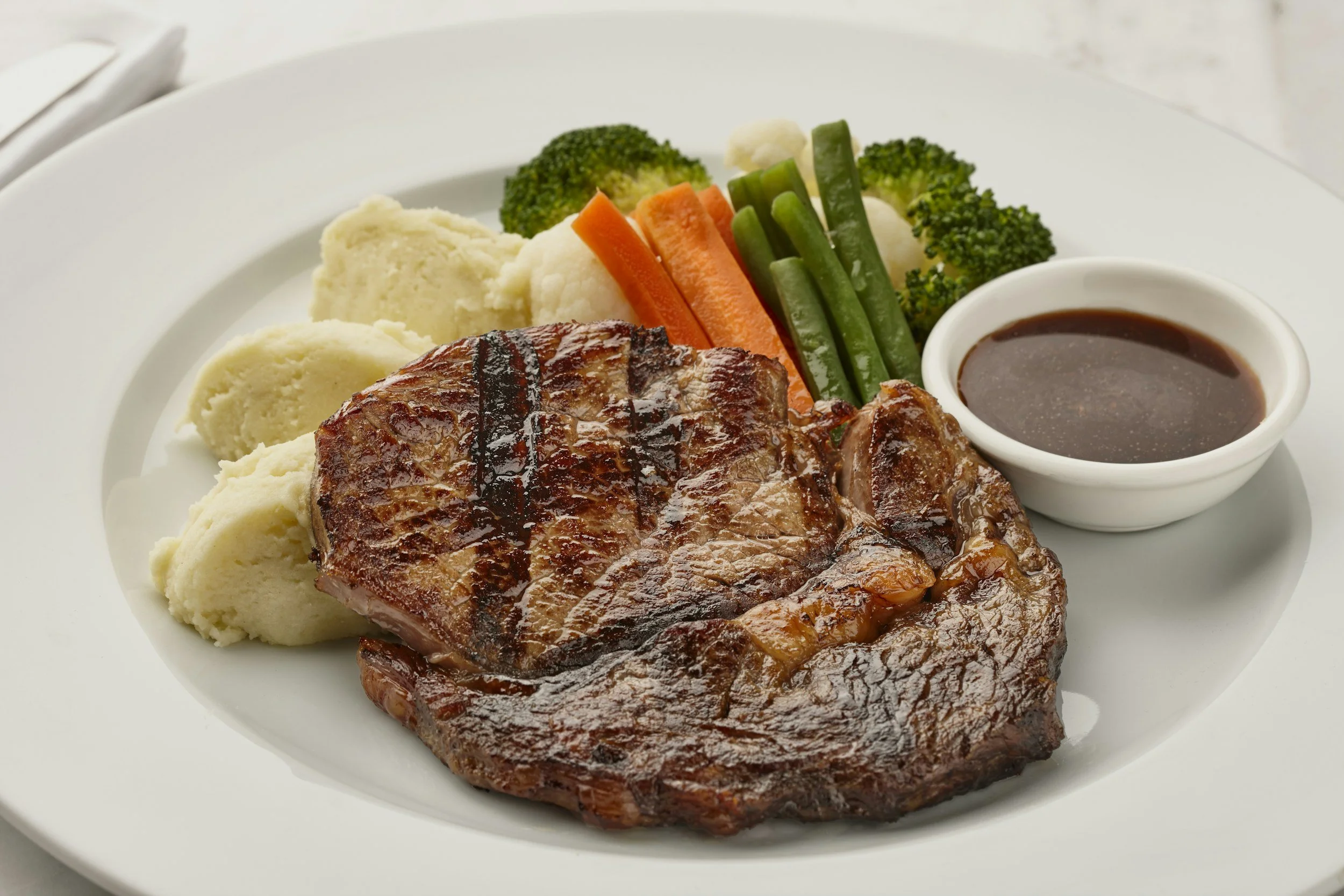 Grilled steak with mashed potatoes, broccoli, cauliflower, carrots, green beans, and a side of sauce.