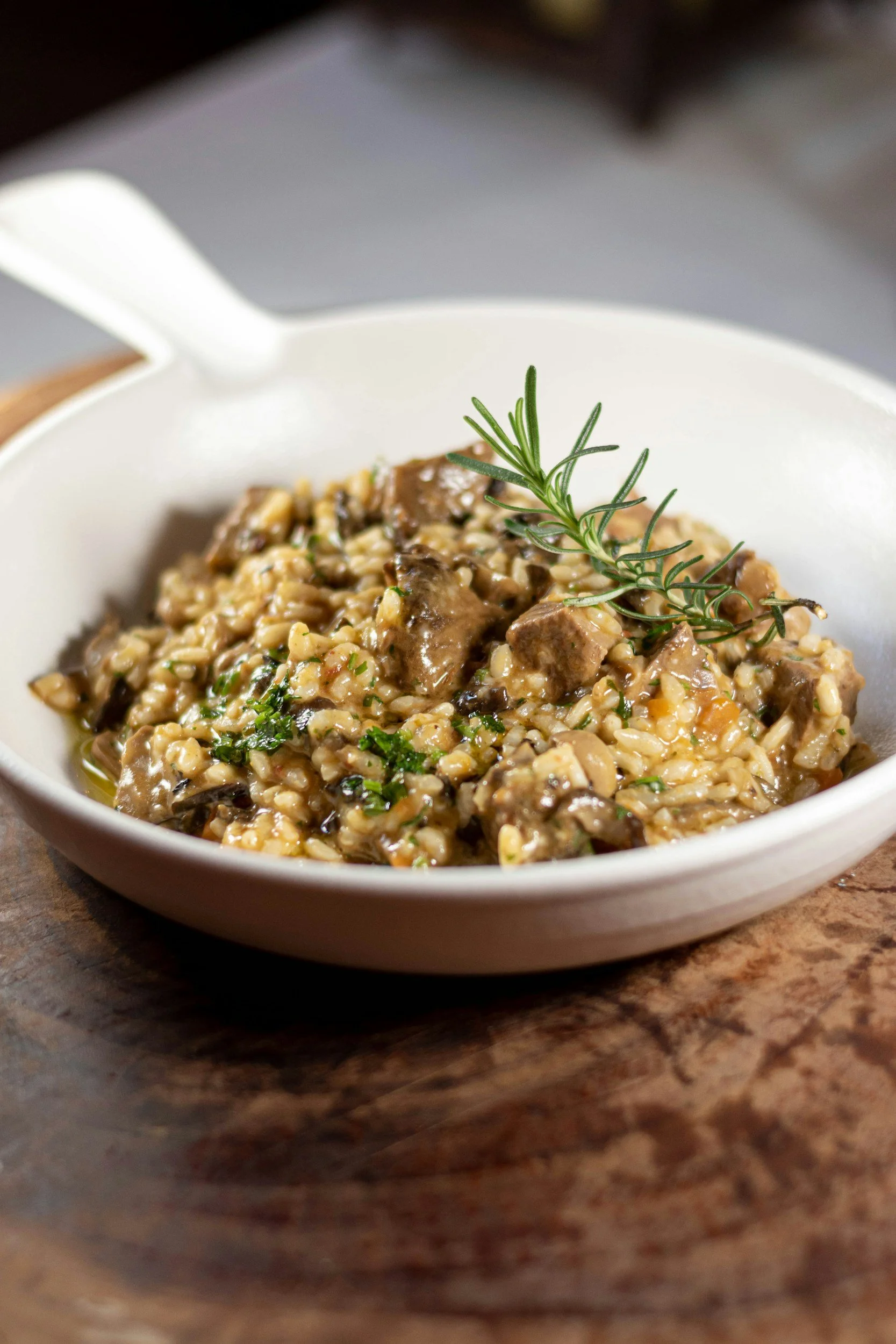 A bowl of beef and rice stew garnished with a sprig of rosemary, on a wooden surface.