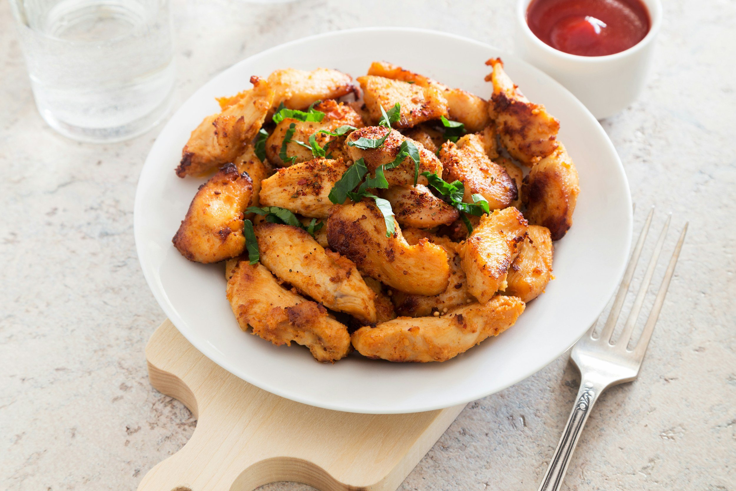 Plate of cooked chicken pieces garnished with herbs, with a glass of water, a small bowl of ketchup, and a fork nearby.