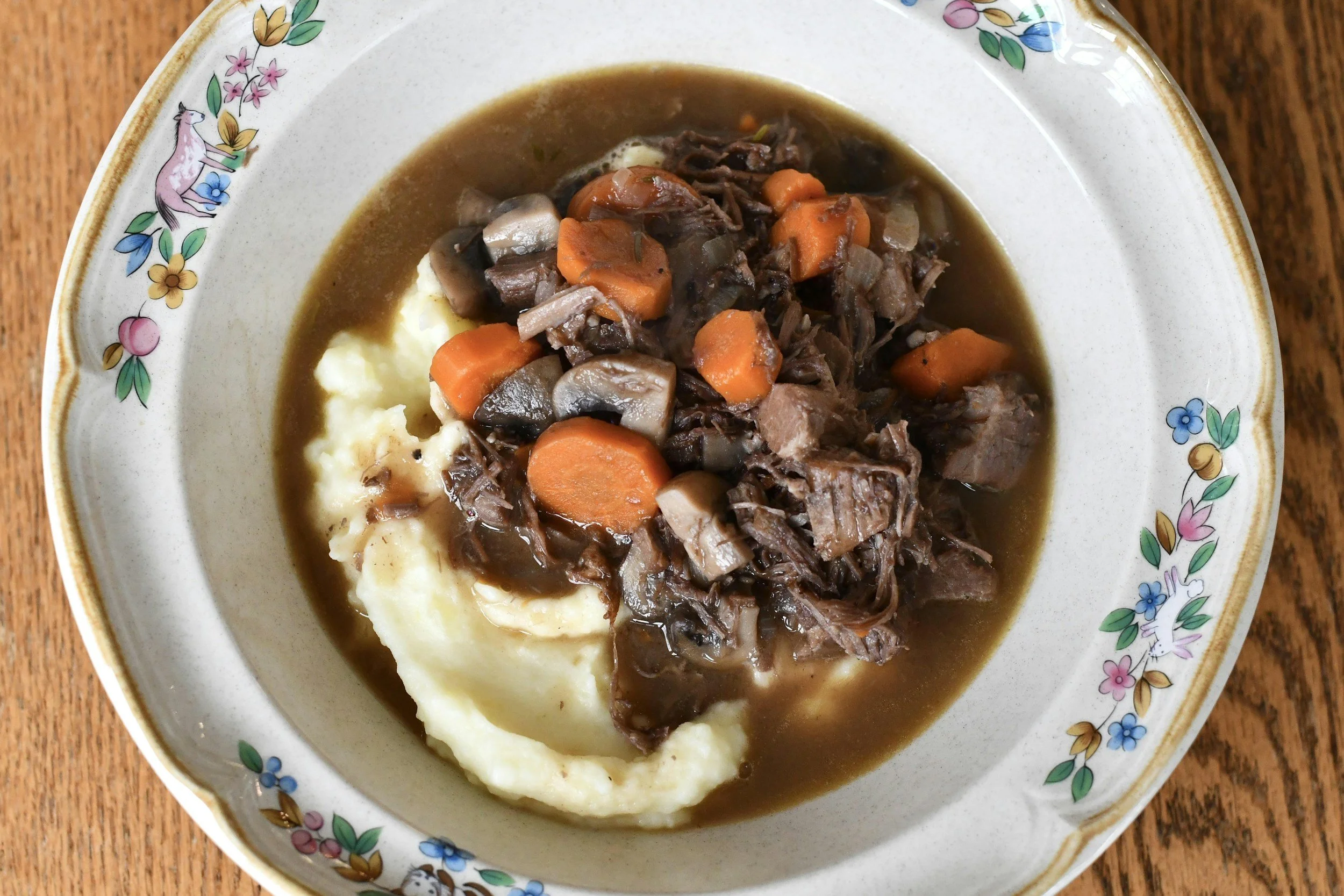 A bowl of beef stew served over mashed potatoes, with chunks of carrots and mushrooms, in a decorative floral china dish.