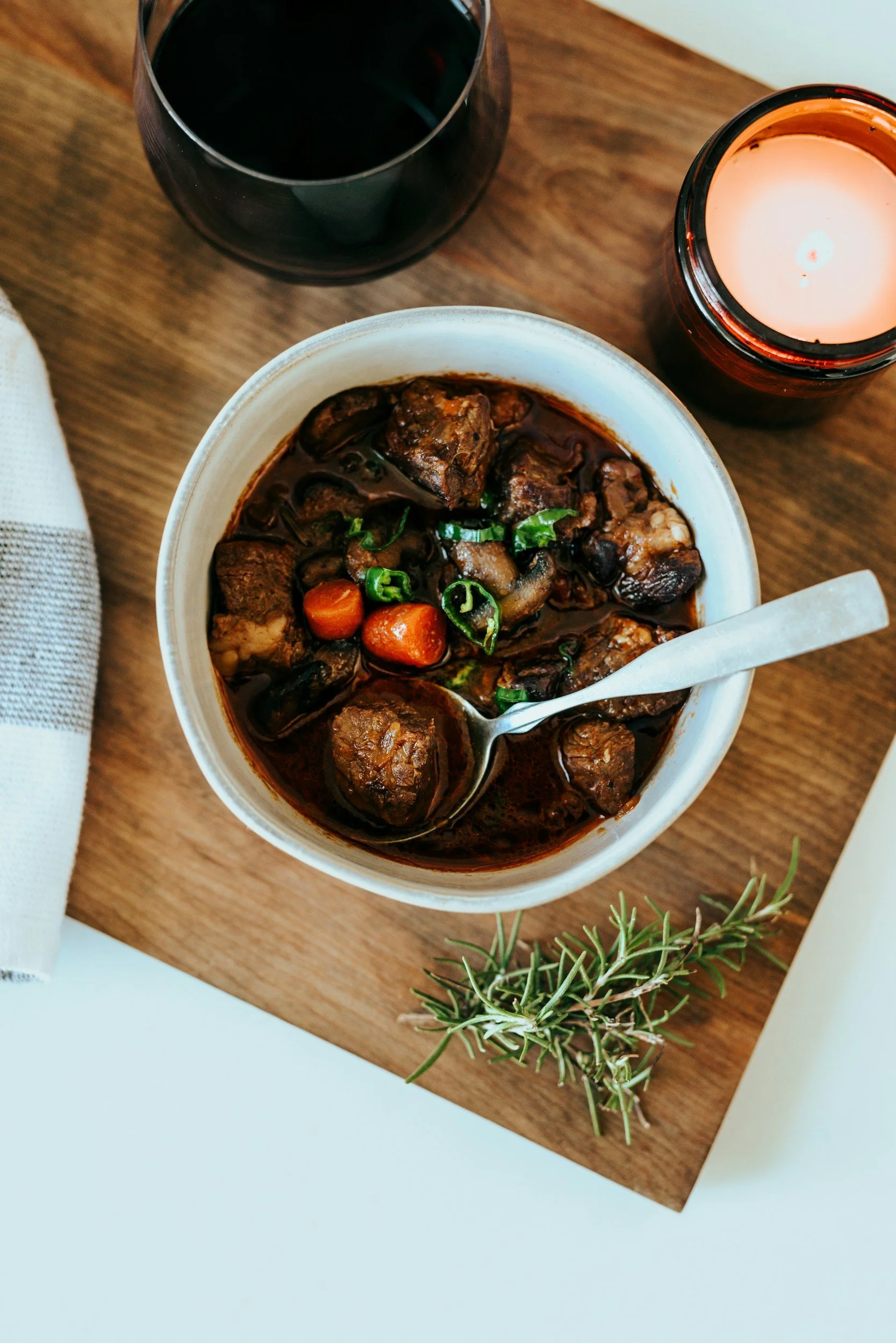 A bowl of beef stew with carrots and green onions, on a wooden tray with a sprig of rosemary, accompanied by a glass of red wine and a lit candle.