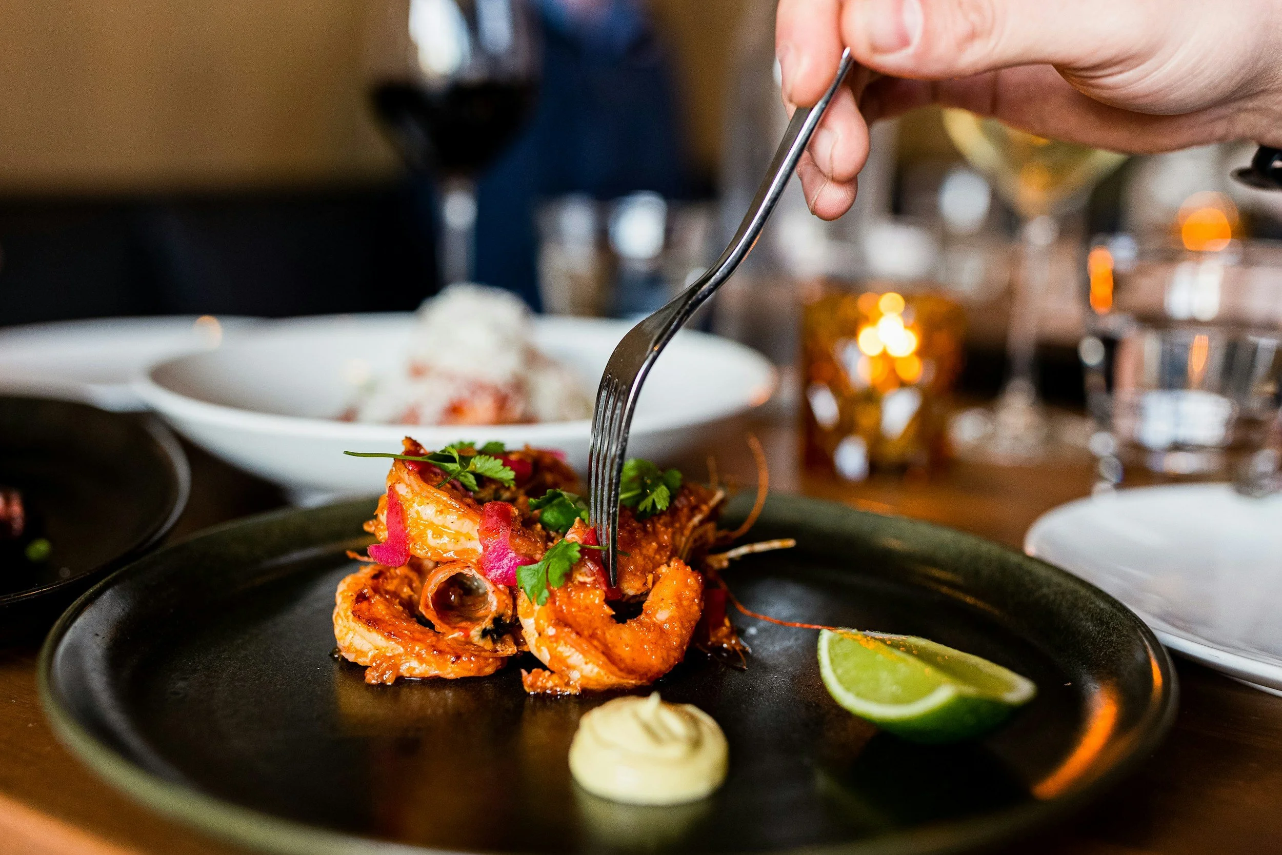 Person using a fork to eat a dish of spicy shrimp garnished with cilantro and pink pickled onions, served with a lime wedge and a dollop of mayonnaise on a black plate, with a glass of red wine in the background.