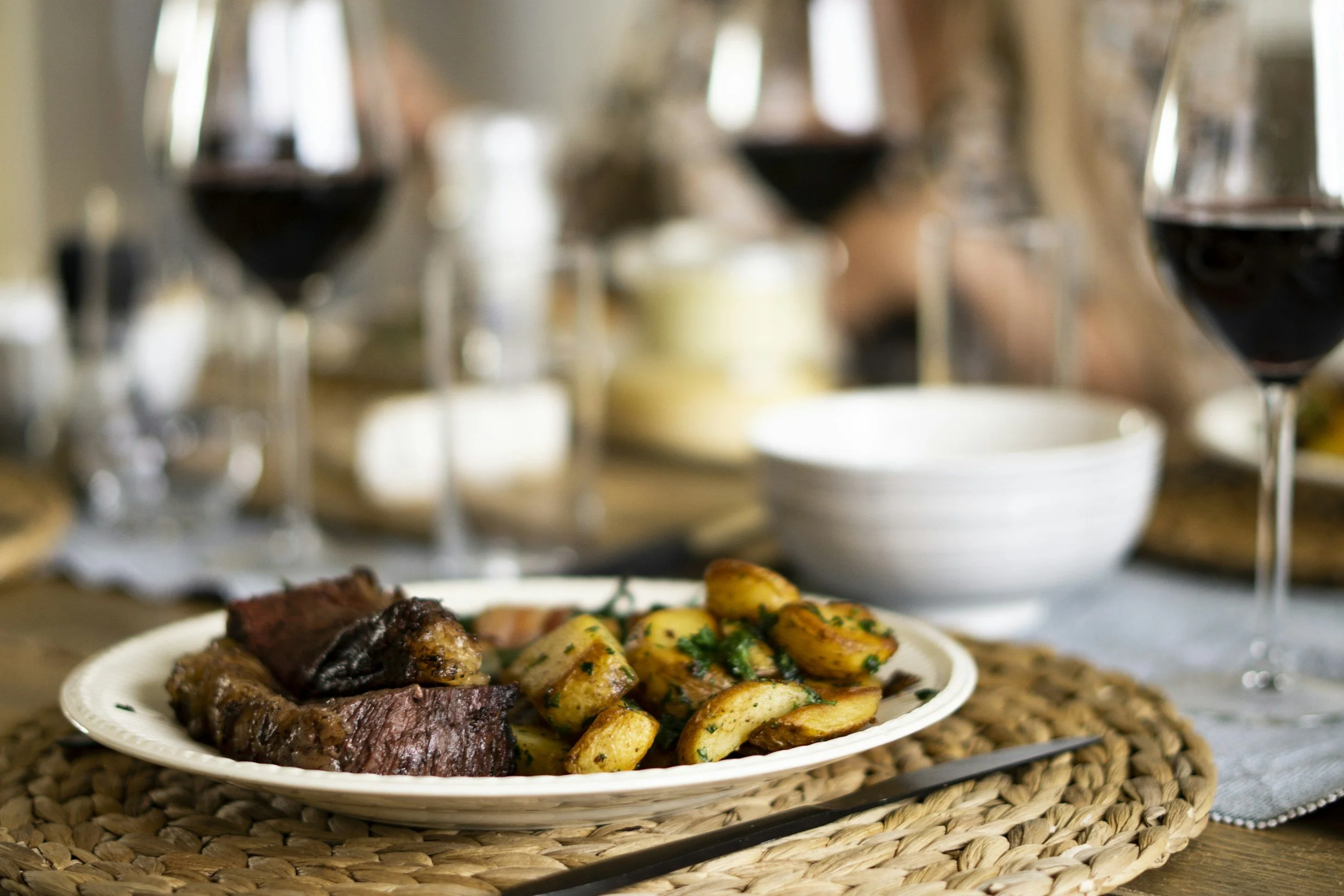 Plate of grilled meat and roasted potatoes on a woven placemat, with glasses of red wine and a bowl in the background.