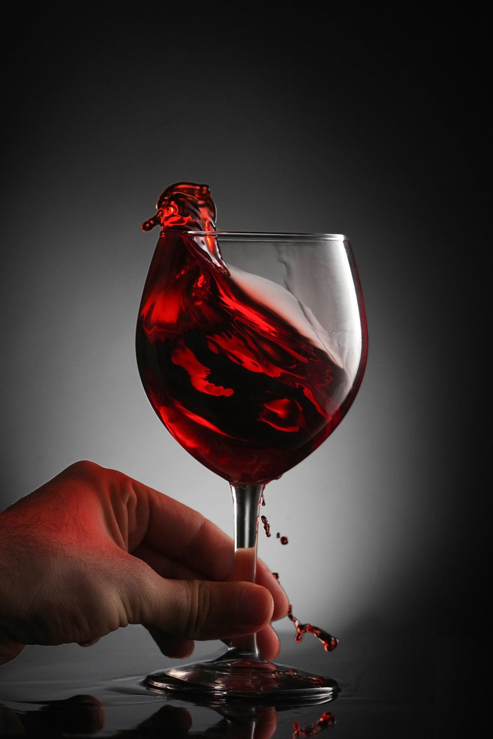 A hand holding a wine glass filled with red wine, captured in motion with some wine spilling over the rim against a dark background.