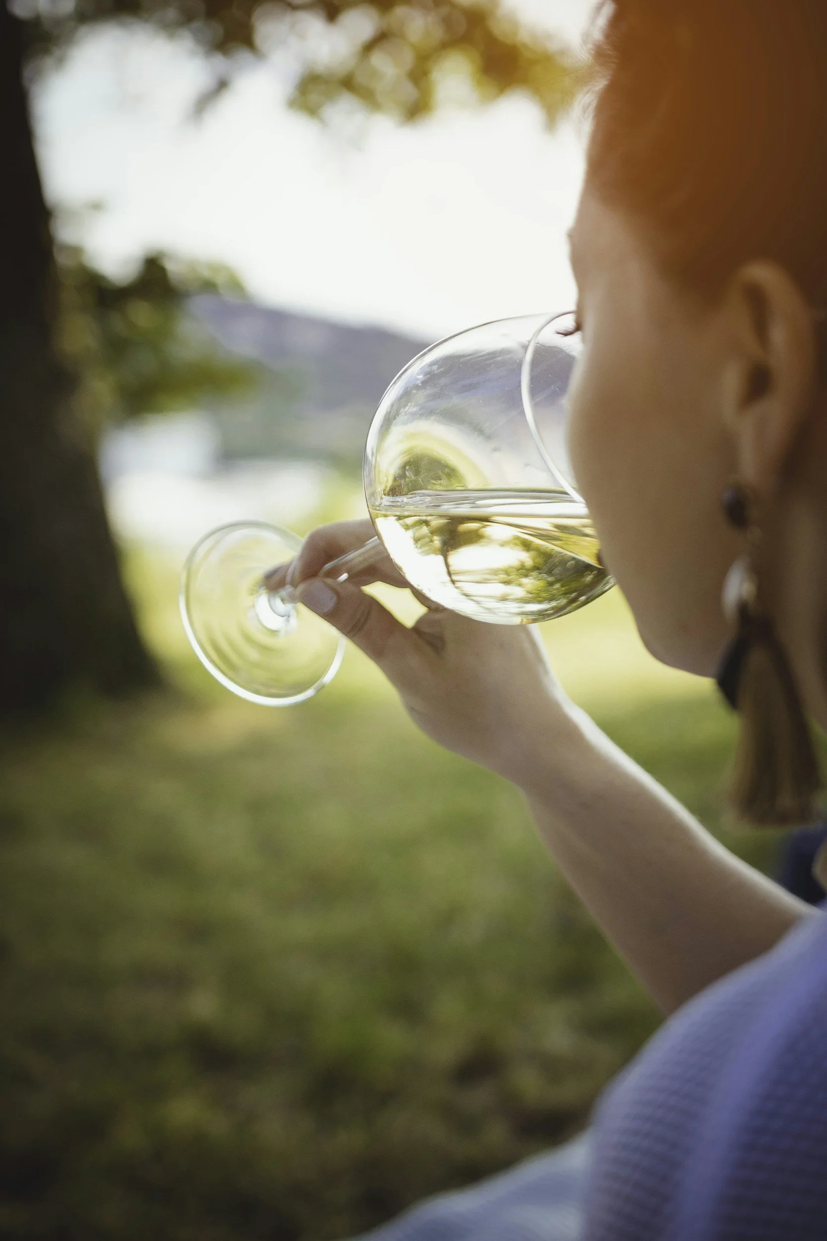 Woman drinking white wine outdoors near trees and mountains in the background.