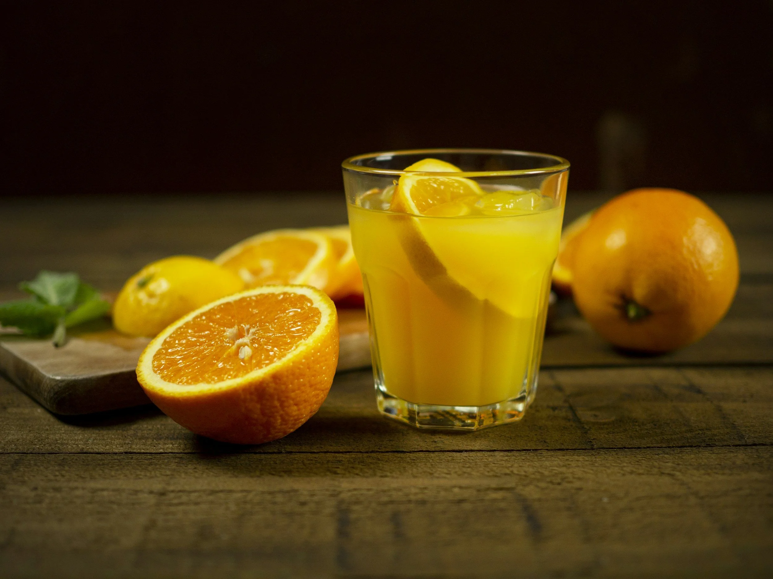A glass of orange juice on a wooden table with oranges and orange slices around it.