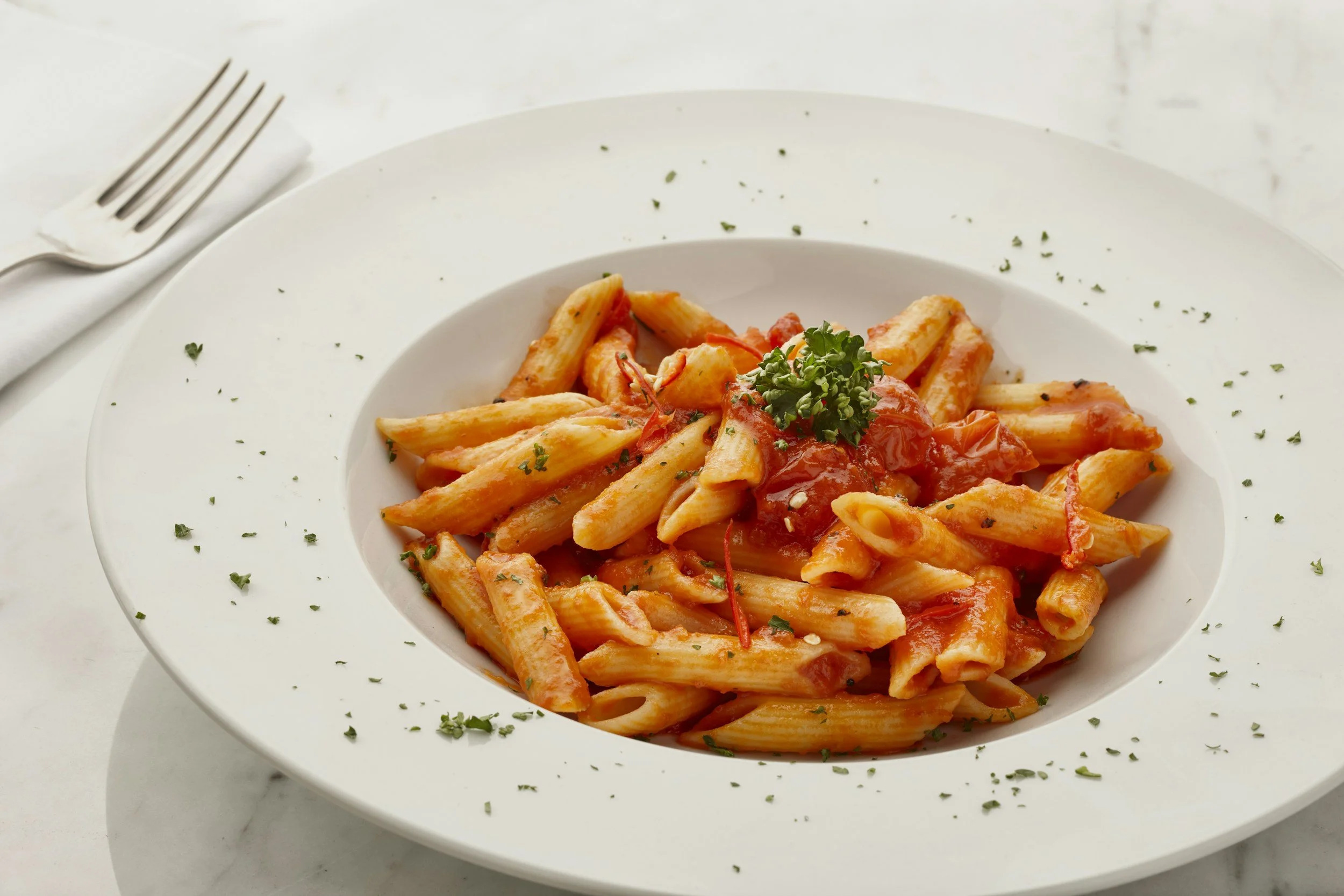 Plate of penne pasta with tomato sauce and garnished with parsley, on a white marble table with a fork and napkin nearby.