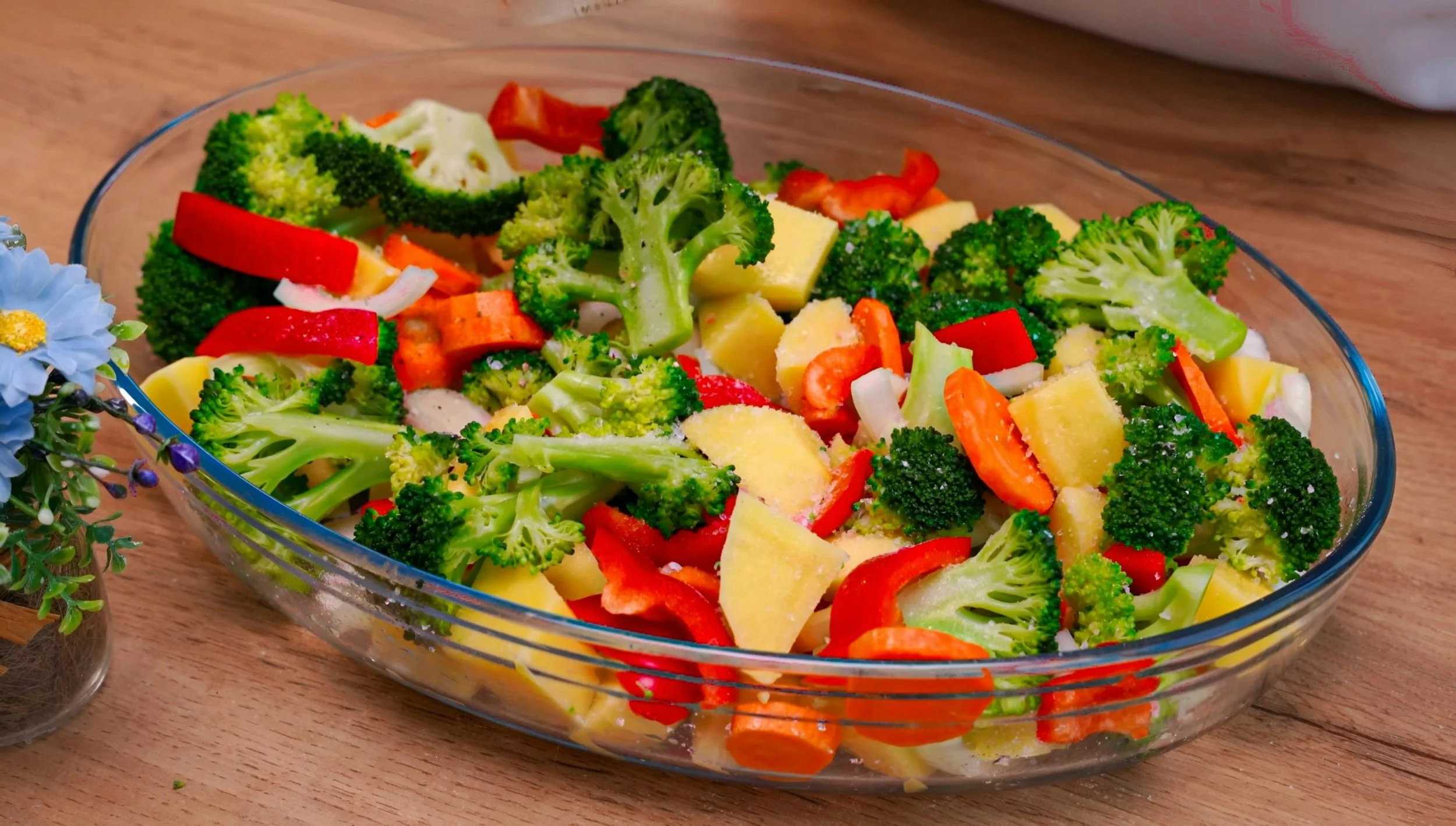 Mixed chopped vegetables in a glass serving dish, including broccoli, red and orange bell peppers, potatoes, and carrots, on a wooden surface.