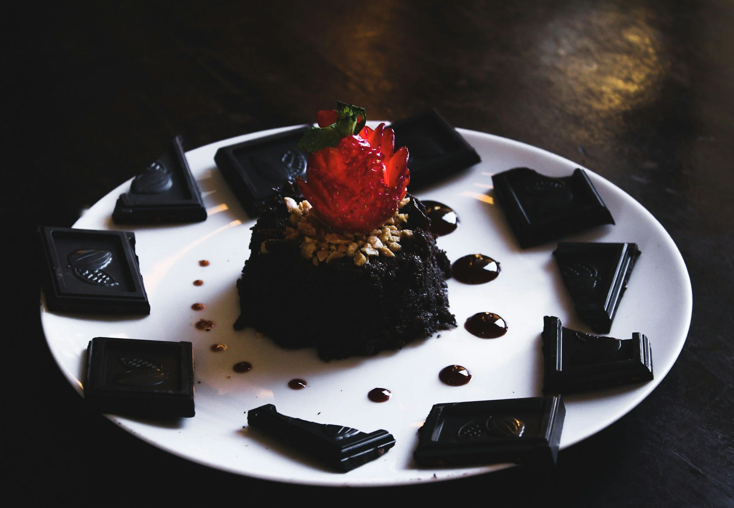 Chocolate dessert topped with a strawberry and crushed nuts, garnished with chocolate sauce on a white plate with chocolate pieces around it.