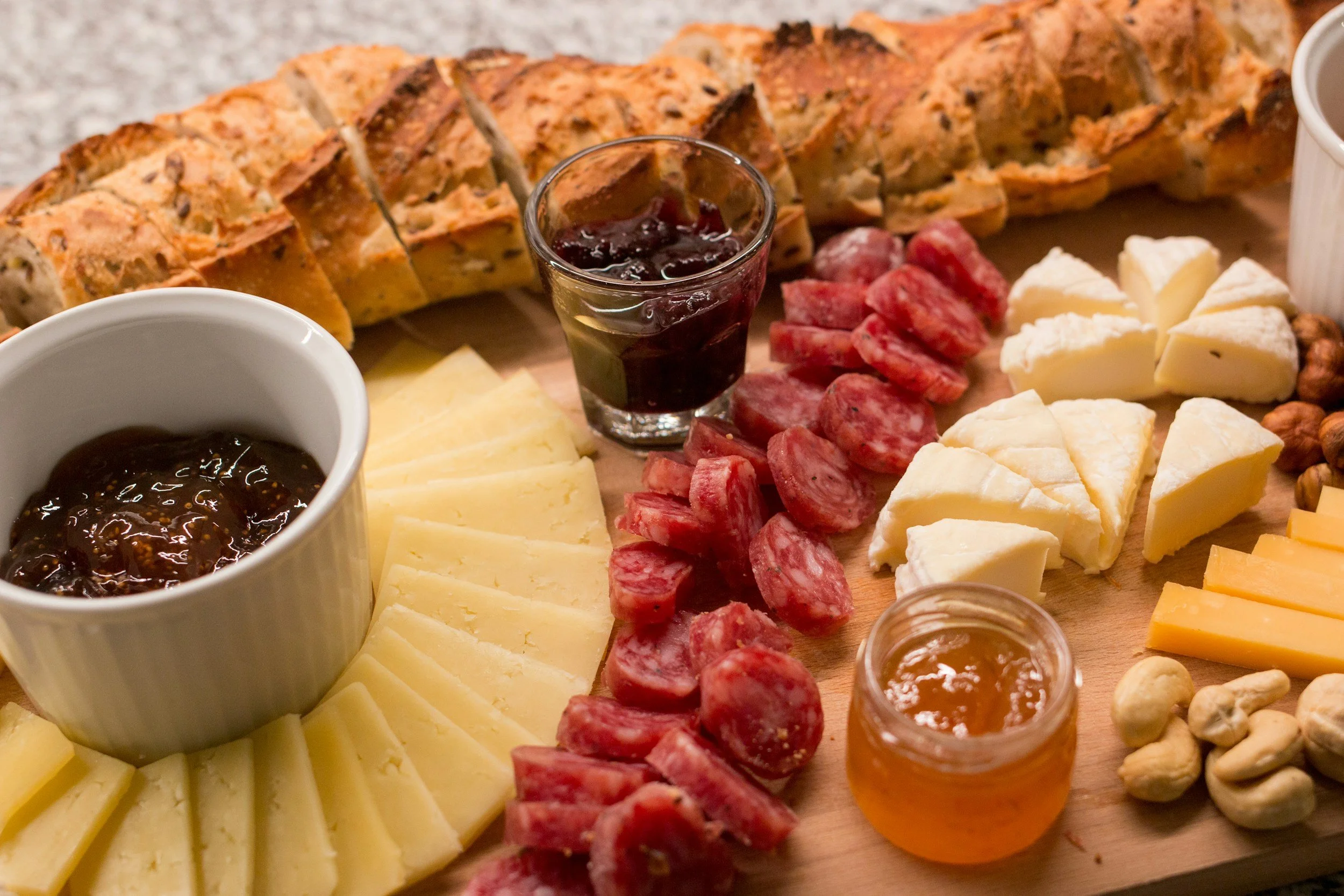 A cheese and charcuterie board with sliced cheese, slices of cured sausage, crackers, jam, honey, nuts, and a small jar of jelly.