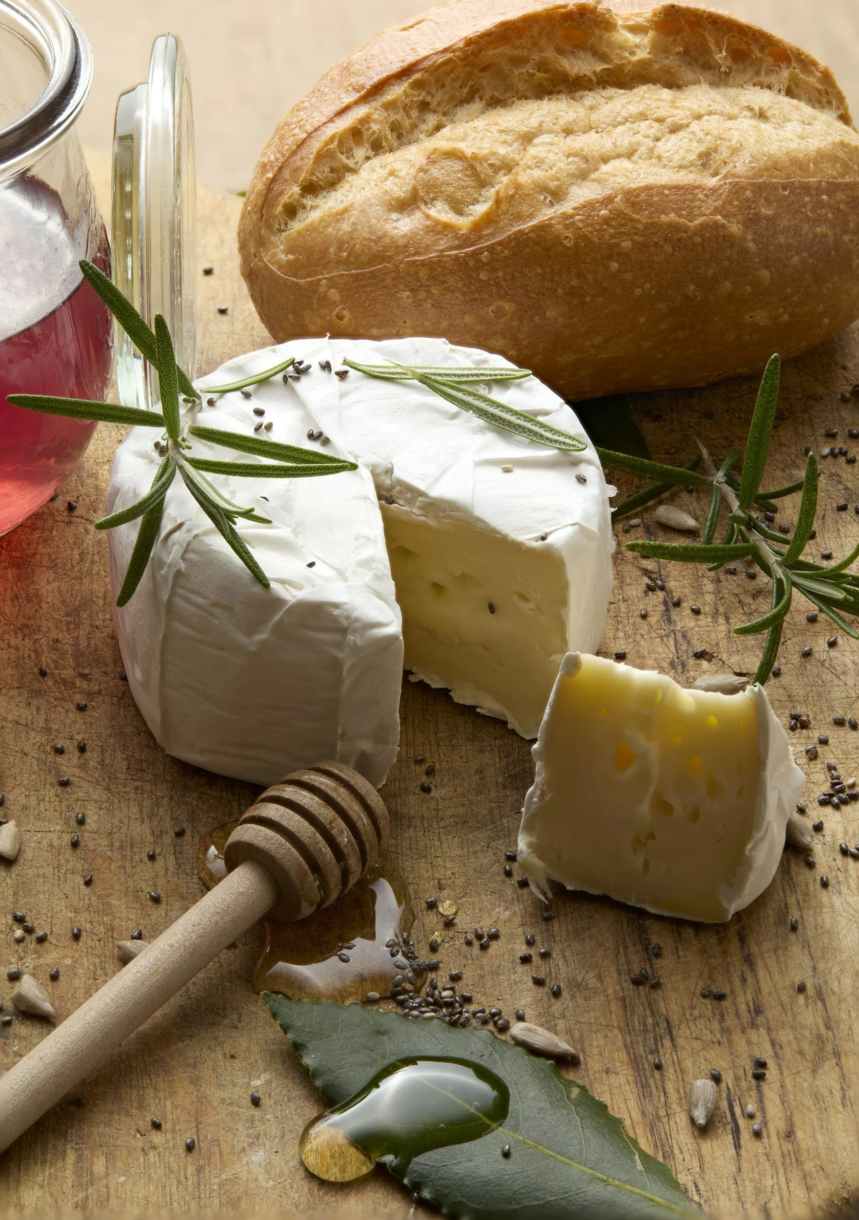 A round wheel of brie cheese garnished with rosemary sprigs, placed on a wooden surface with scattered black peppercorns, sunflower seeds, and honey, alongside a chopped piece of cheese, a loaf of bread, and a jar of red wine.
