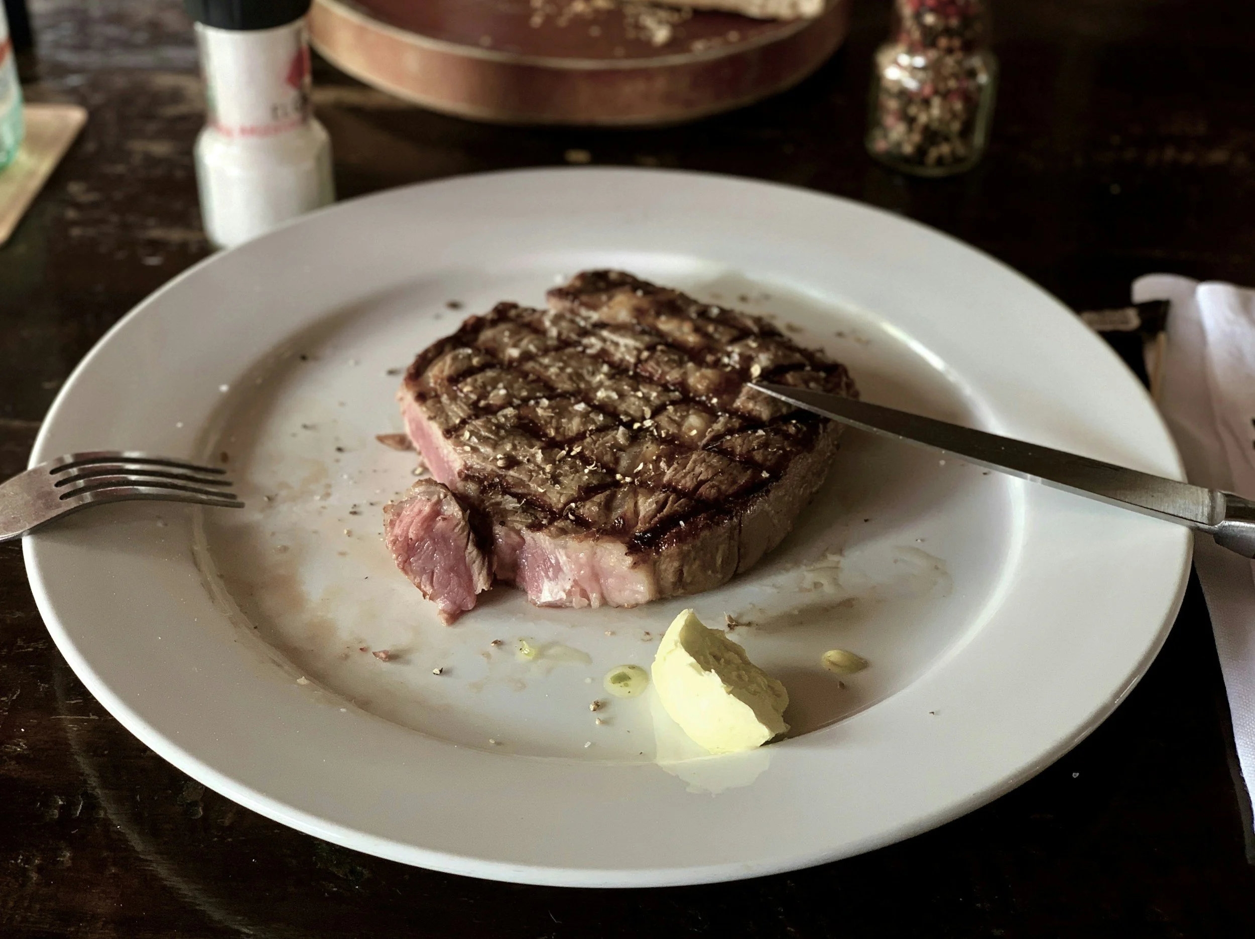 A partially eaten grilled steak on a white plate with a small dollop of butter and some seasoning, on a dark wooden table.
