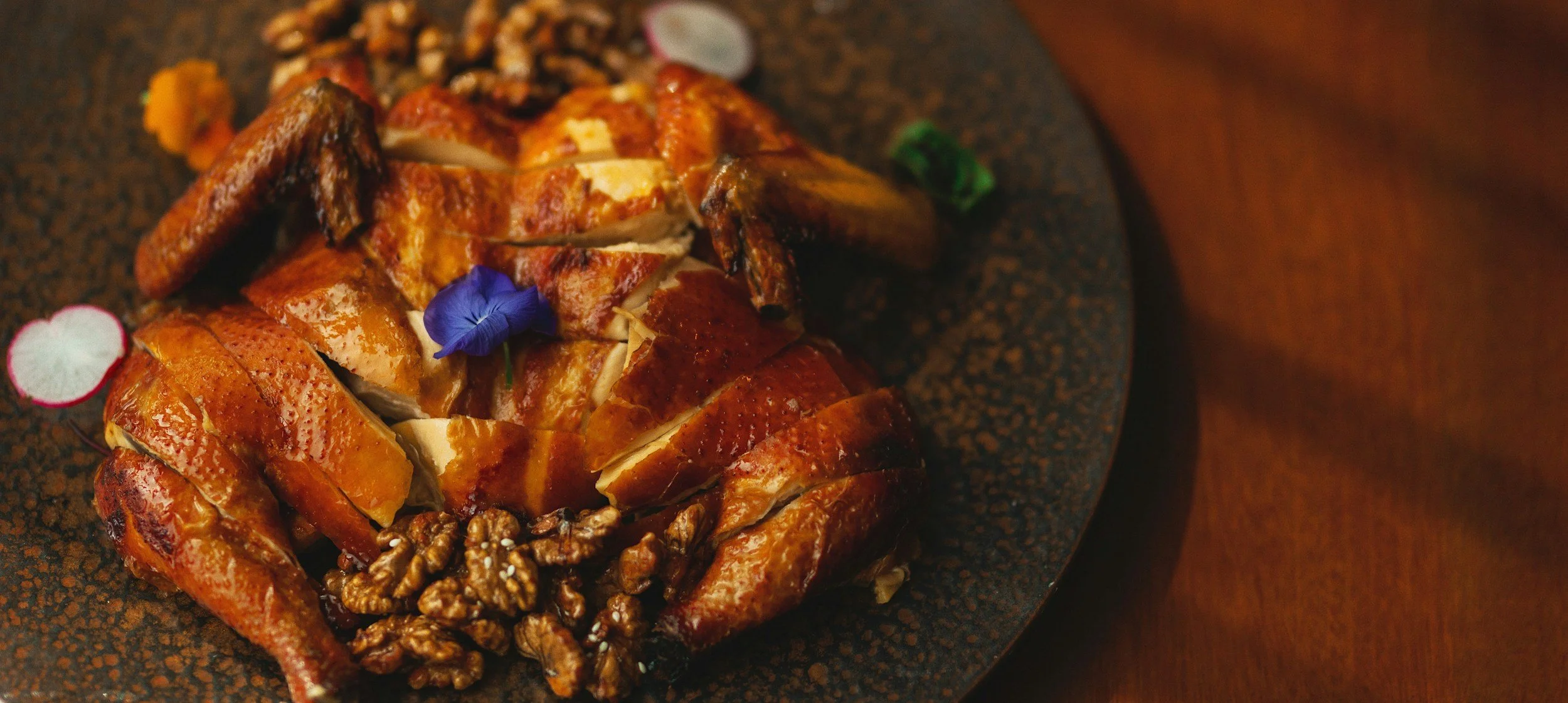 A roasted whole chicken garnished with edible flowers and slices of radish, served on a dark textured plate.