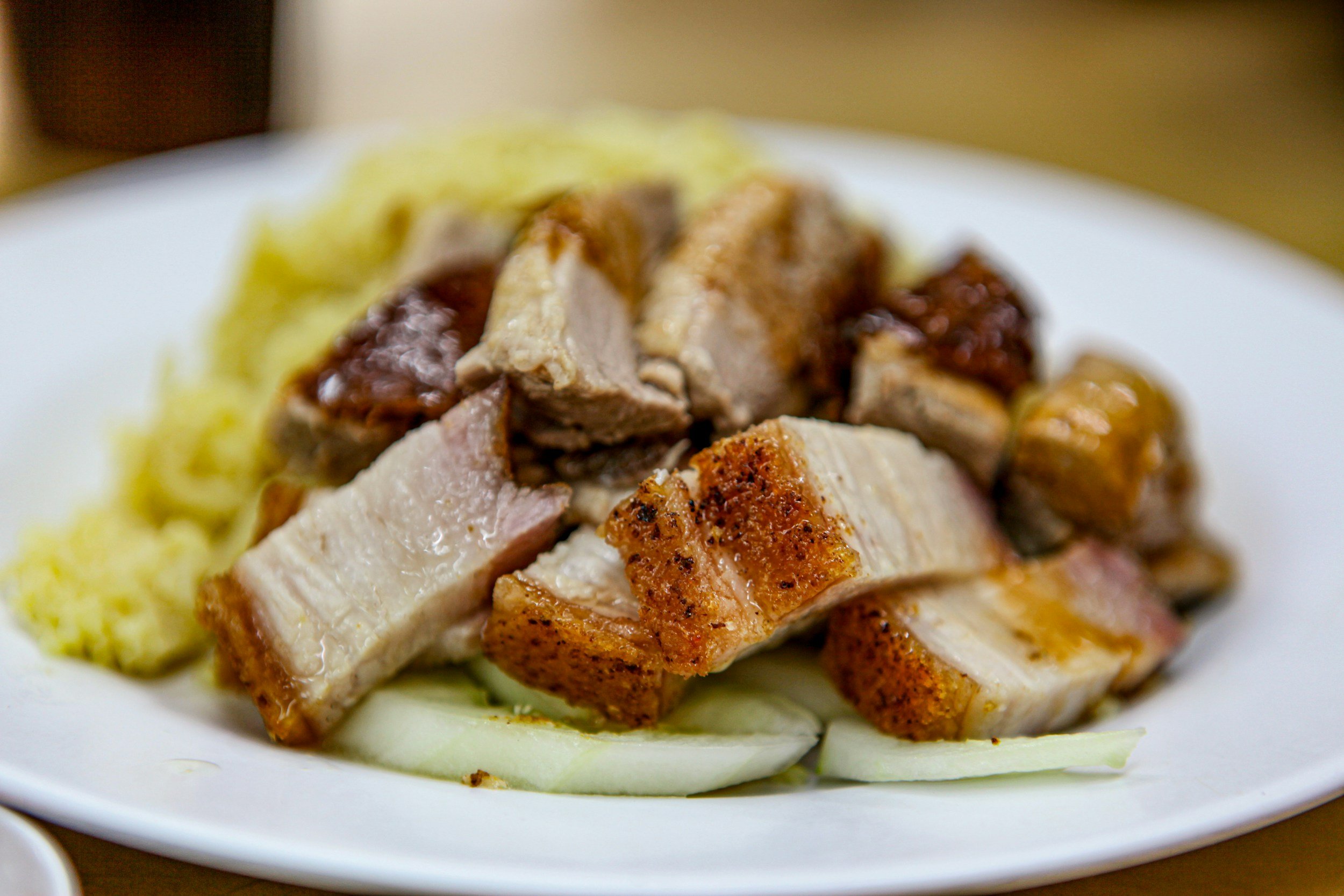 A plate of sliced roasted pork with crispy skin, served on a bed of sliced cucumber, accompanied by mashed potatoes.