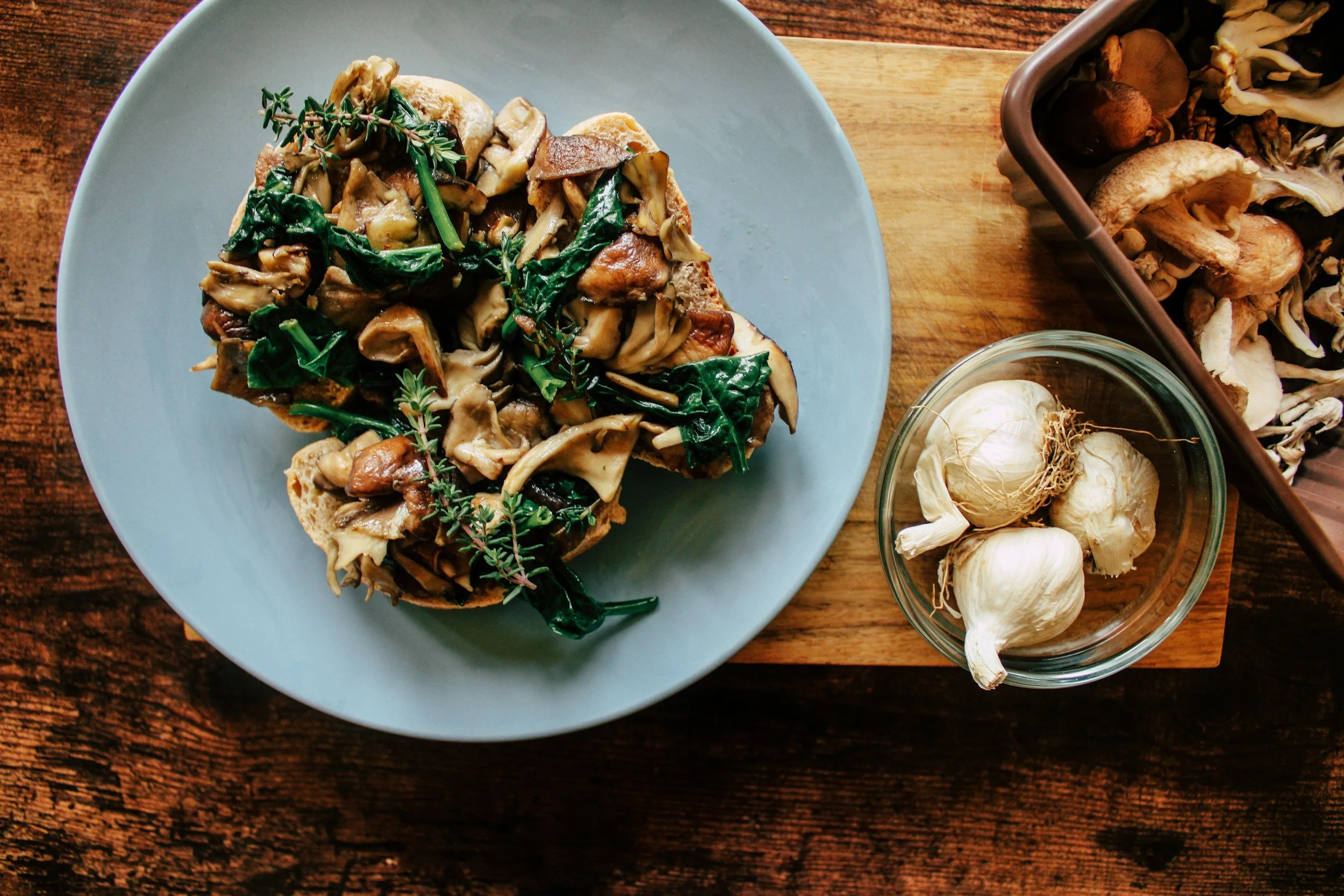 A plate of toasted bread topped with sautéed mixed mushrooms and fresh herbs, with a small glass bowl of garlic cloves and a tray of more mushrooms on a wooden surface.