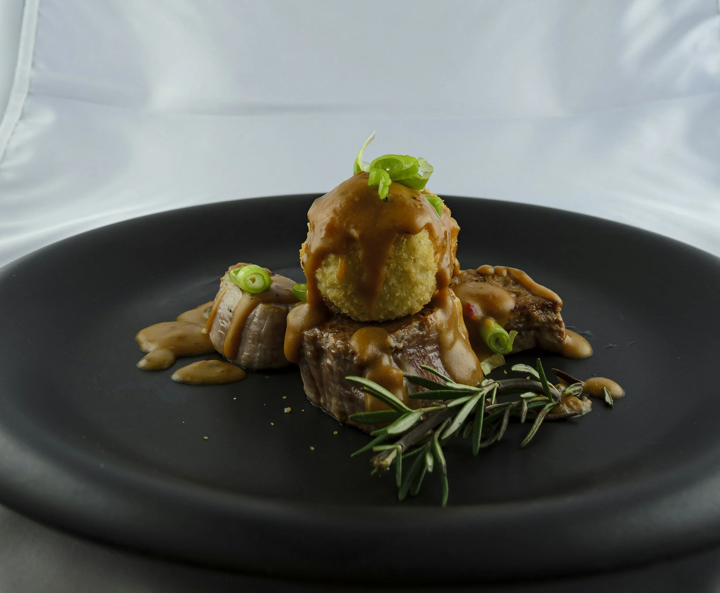 A plated dish with meat, a vegetable topped with sauce, garnished with green onions and rosemary on a black plate.