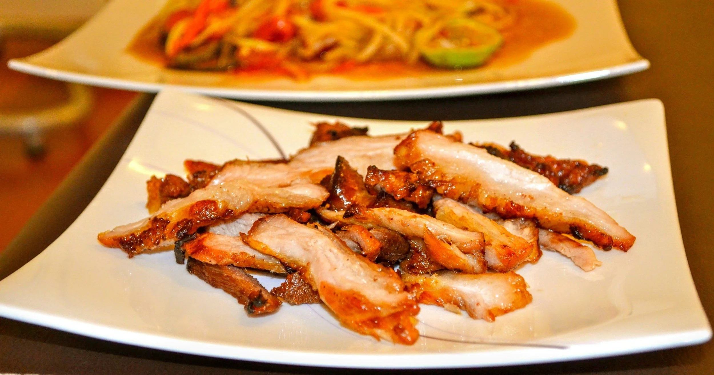 Slices of grilled or roasted meat on a white square plate with another plate of Asian noodle dish in the background