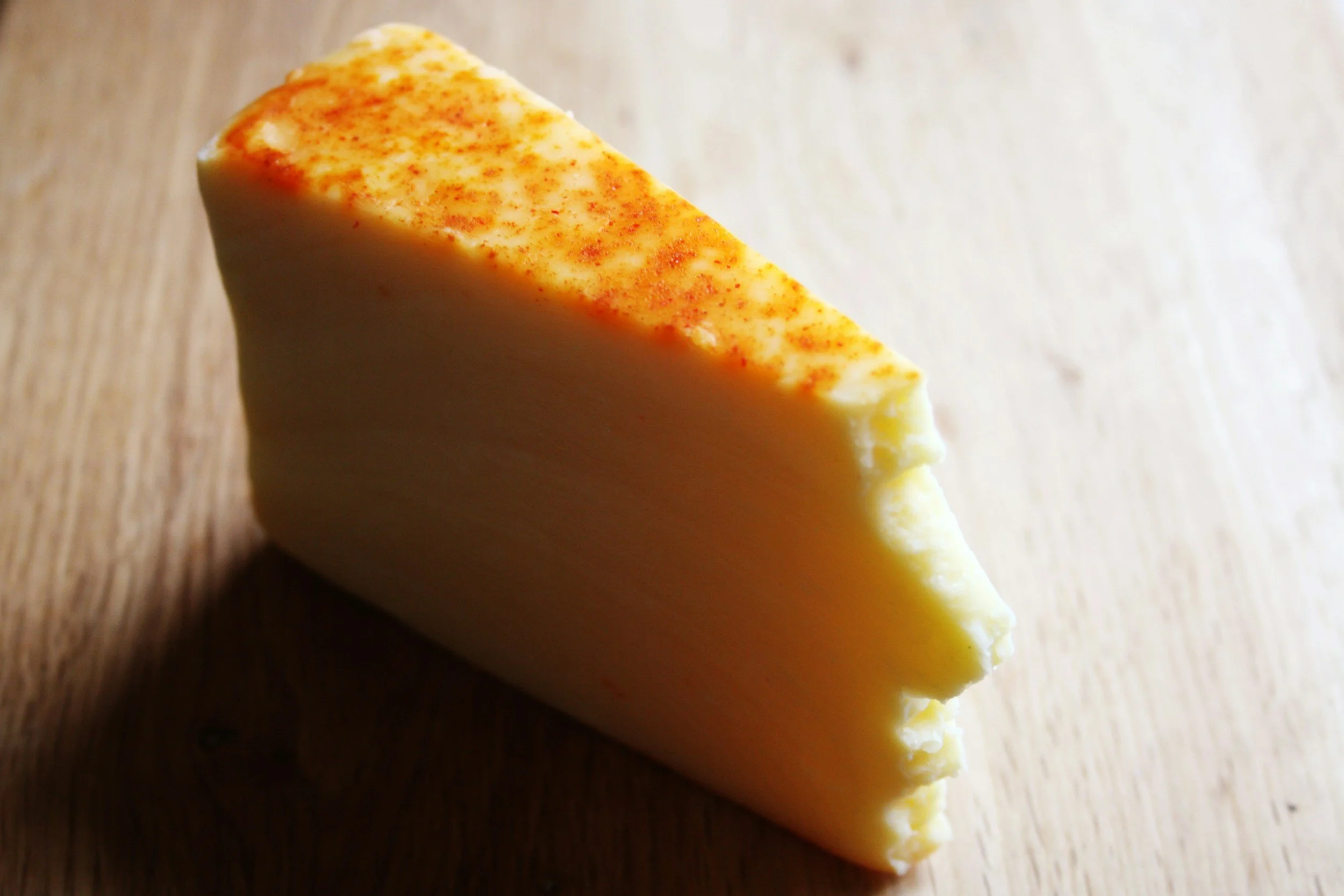 A wedge of semi-hard cheese with a reddish-orange sprinkle on top, placed on a wooden surface.