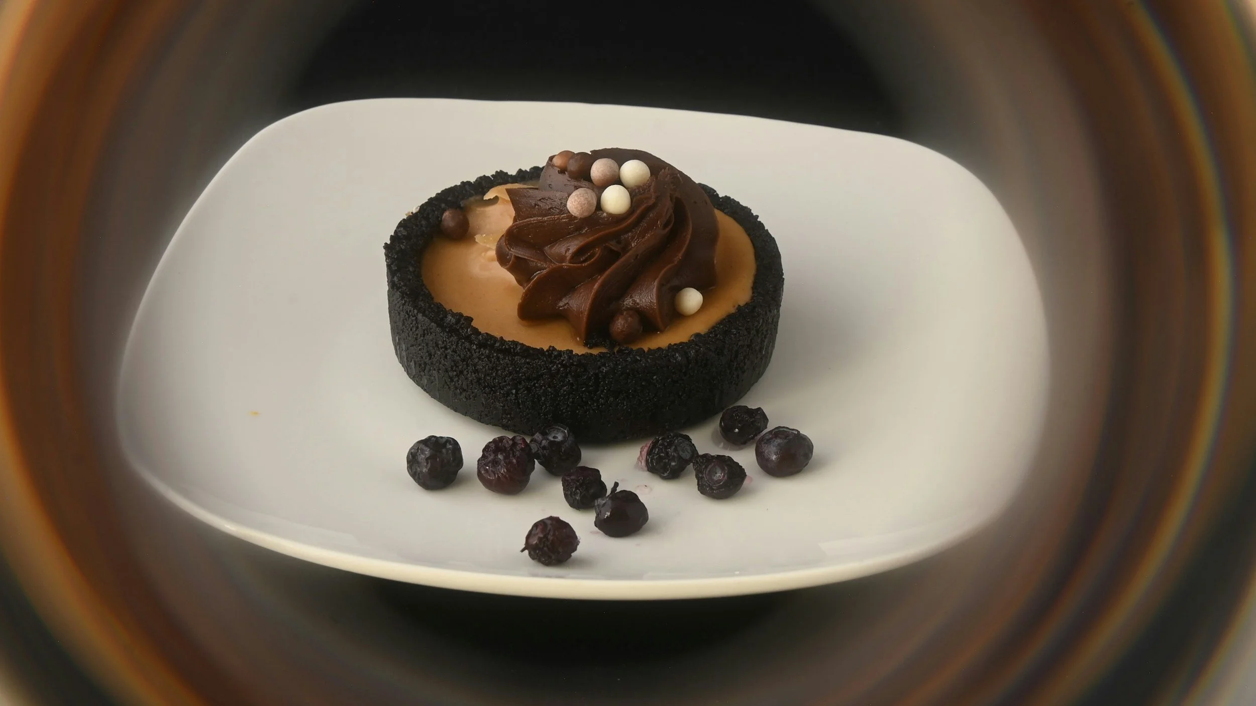A dessert with a black outer crust, filled with caramel, topped with chocolate swirls and small white and brown spherical sprinkles, and garnished with scattered blueberries on a white plate.