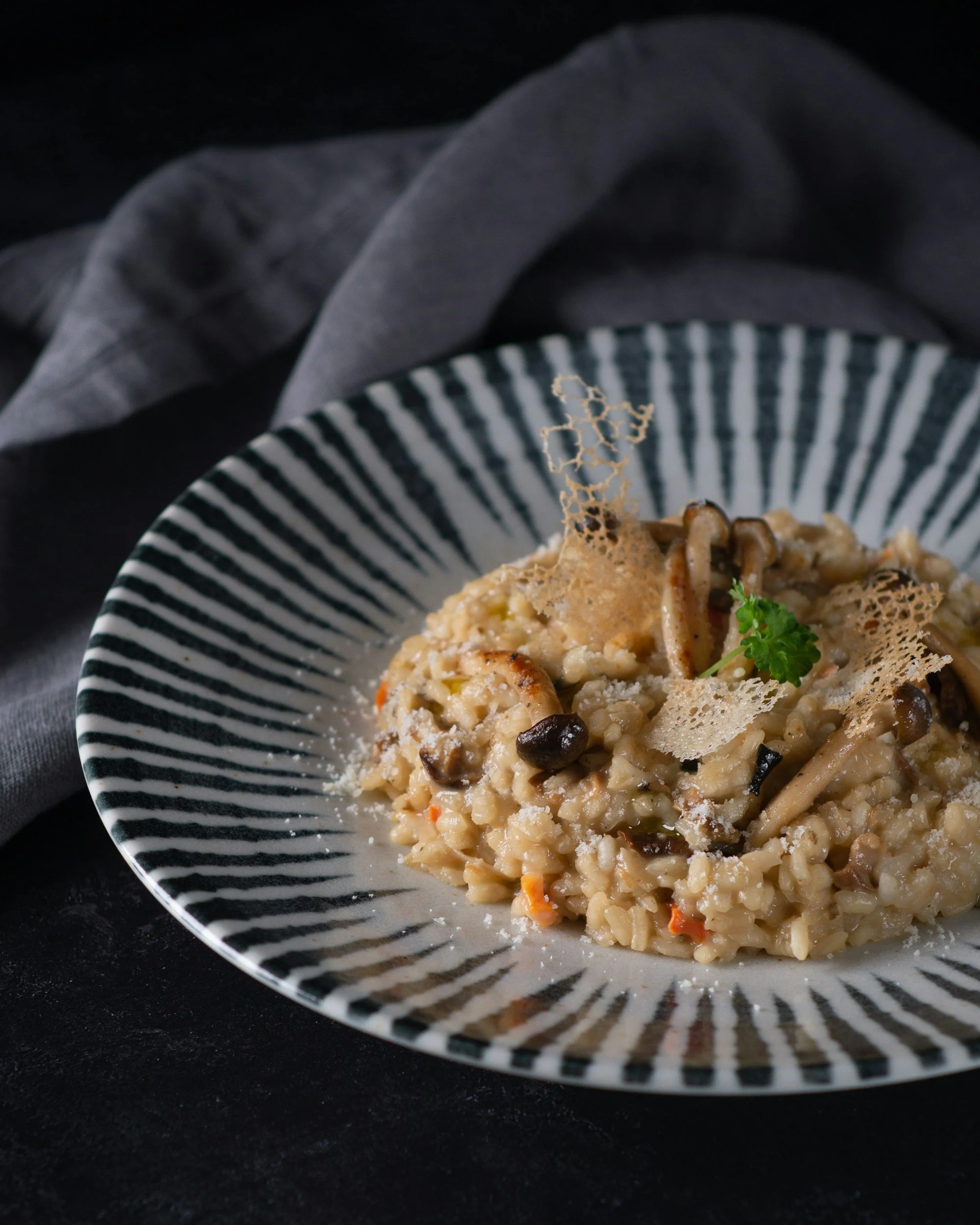 A bowl of mushroom risotto garnished with a sprig of parsley, parmesan cheese, and garnished with a cheese crisp, on a dark surface with a dark cloth in the background.