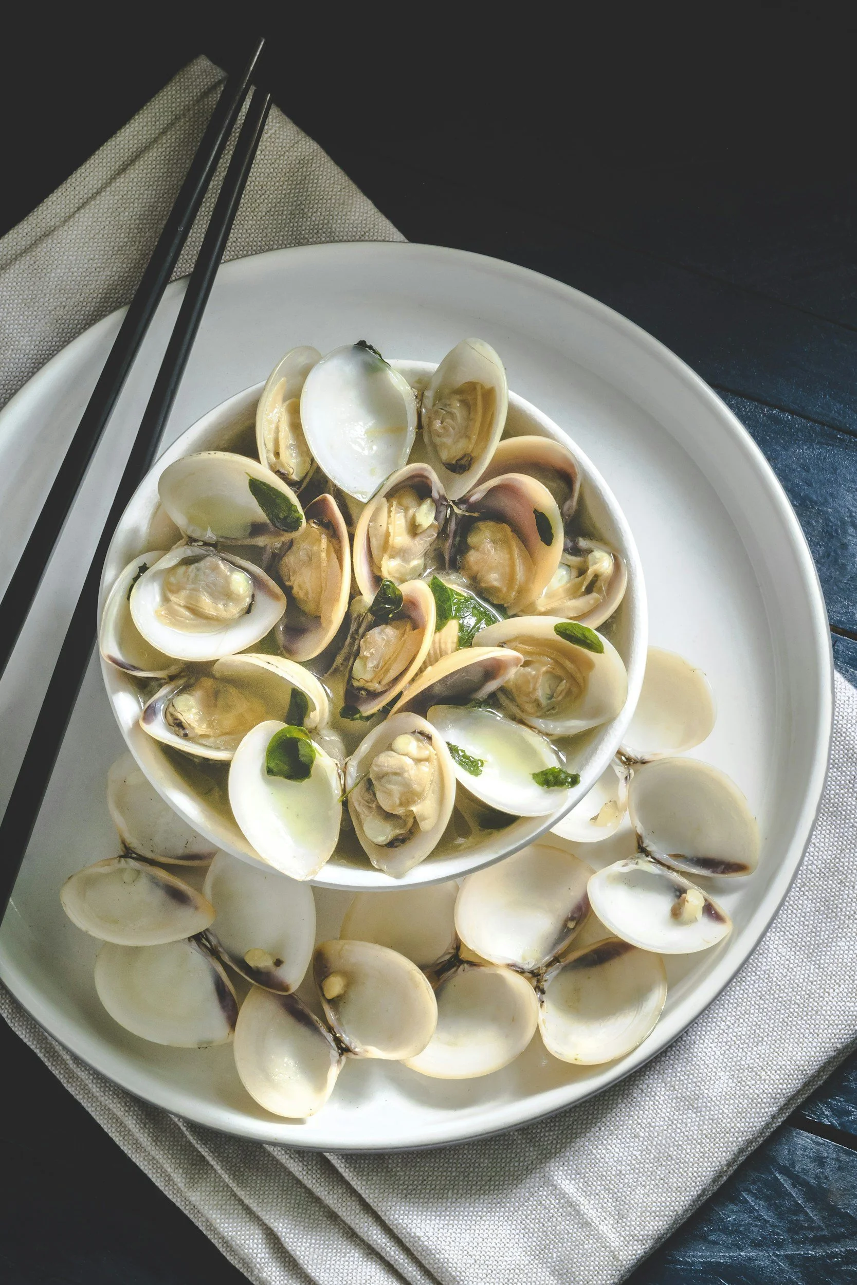 Bowl of cooked clams with green herbs, served on a large white platter with chopsticks.