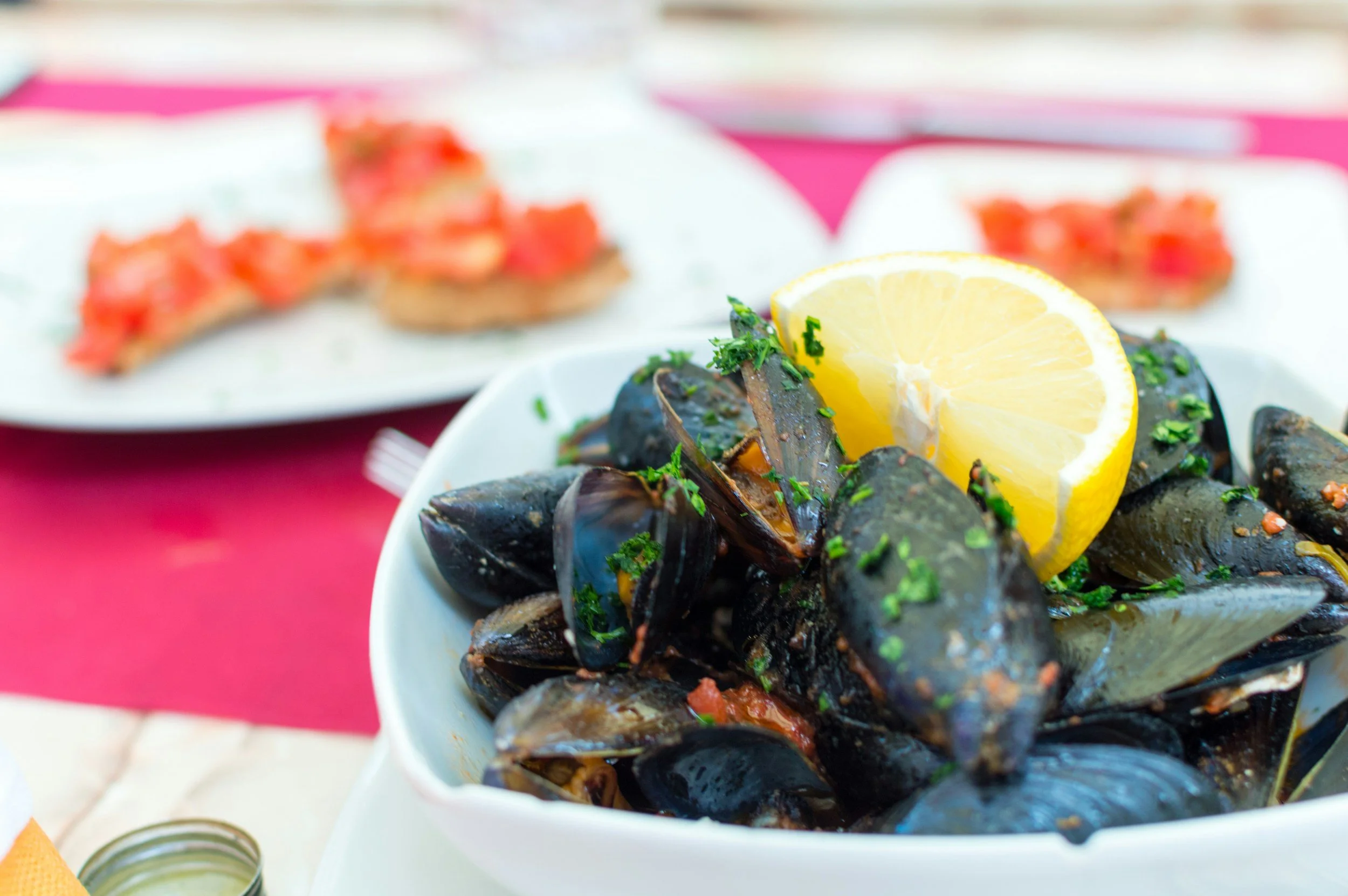 Bowl of cooked mussels garnished with chopped herbs and lemon wedges, with a blurred plate of bread topped with tomato in the background.