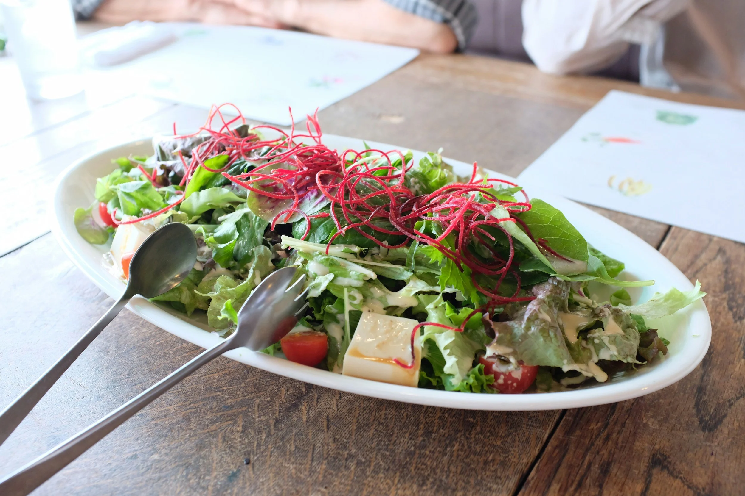 Fresh mixed green salad with cherry tomatoes, cheese cubes, and shredded beets on a white oval plate, on a wooden table.