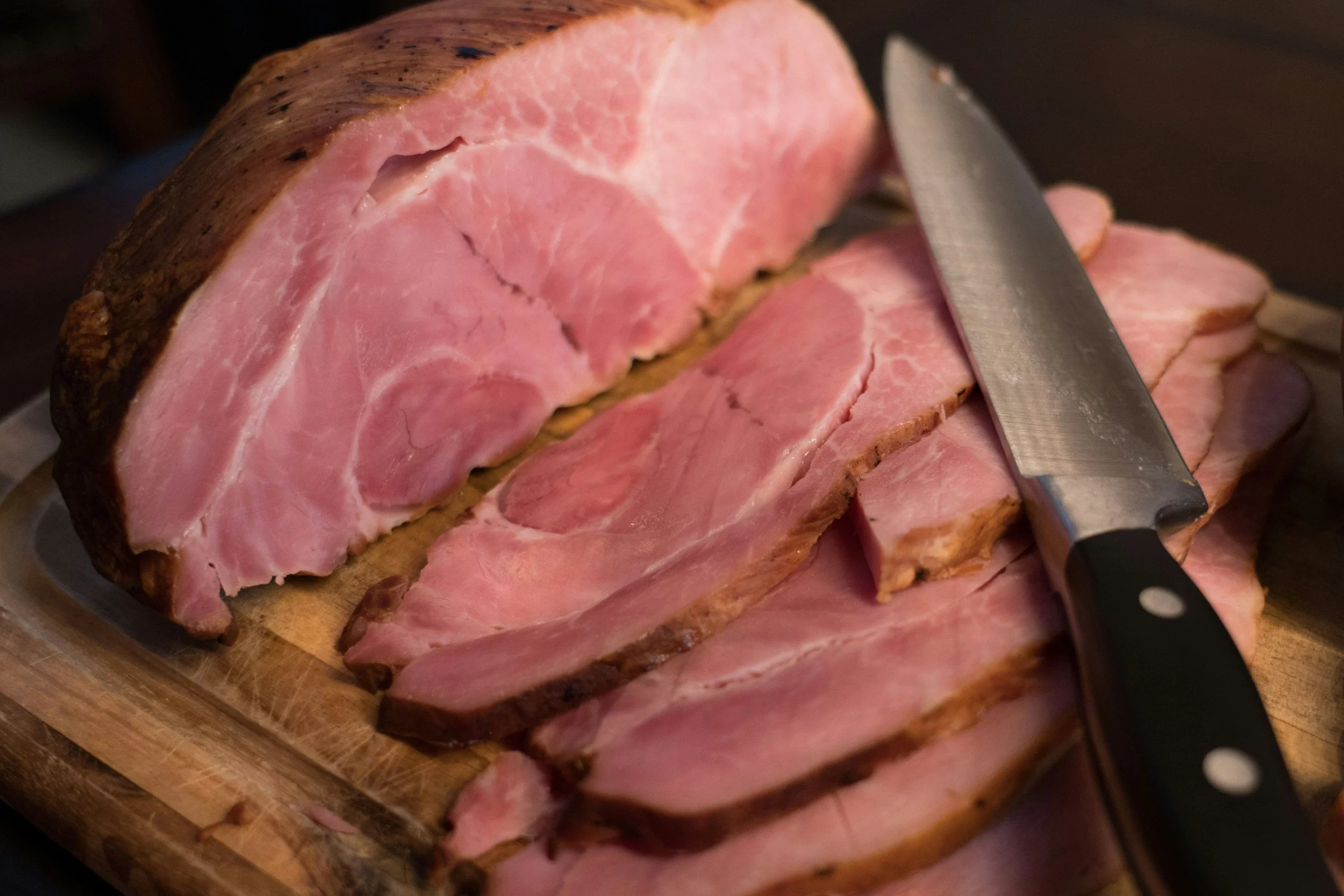 Sliced ham on a wooden cutting board with a large knife resting on top.
