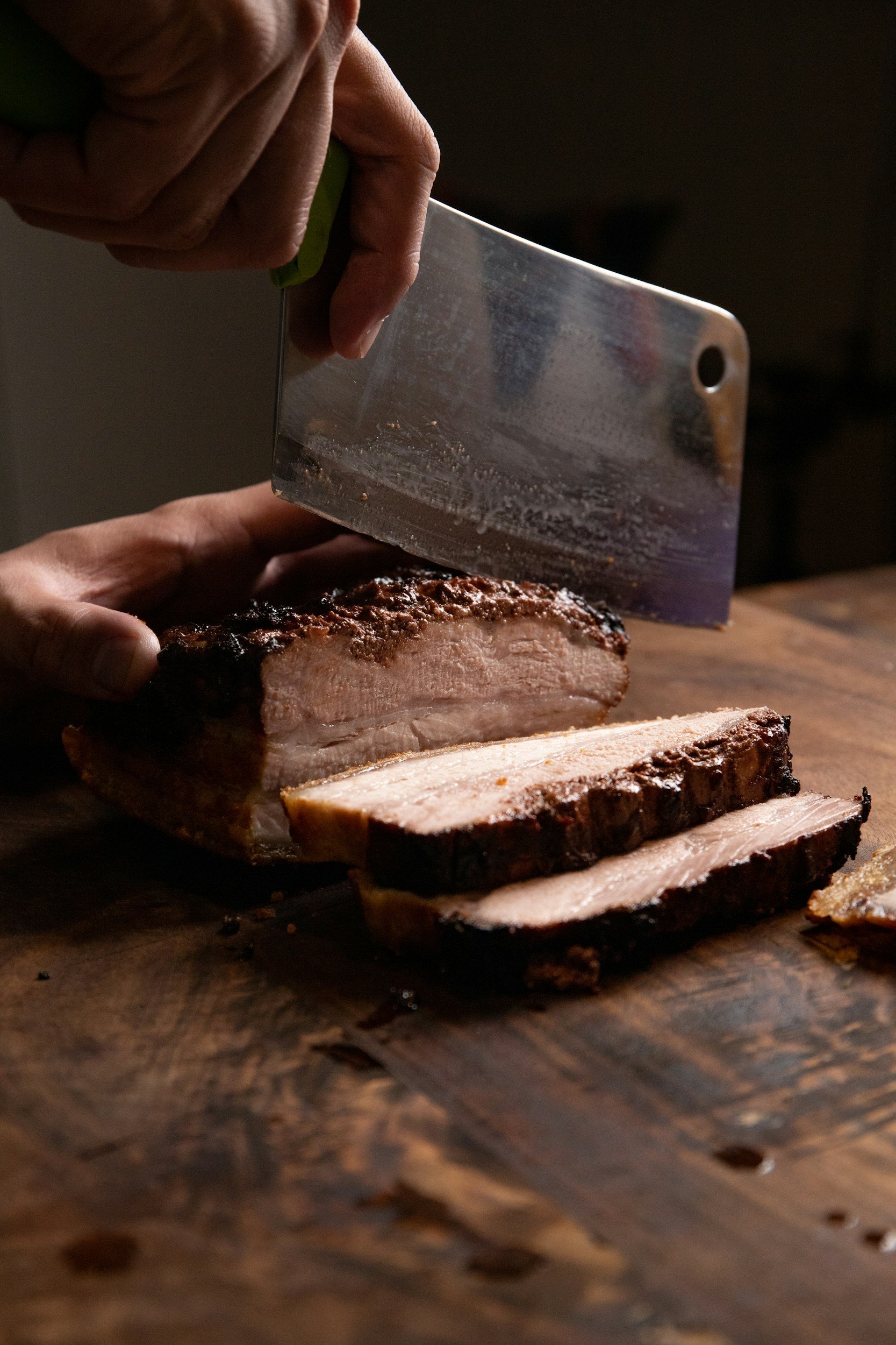 Person slicing a cooked piece of meat, possibly brisket, on a wooden cutting board with a large knife.