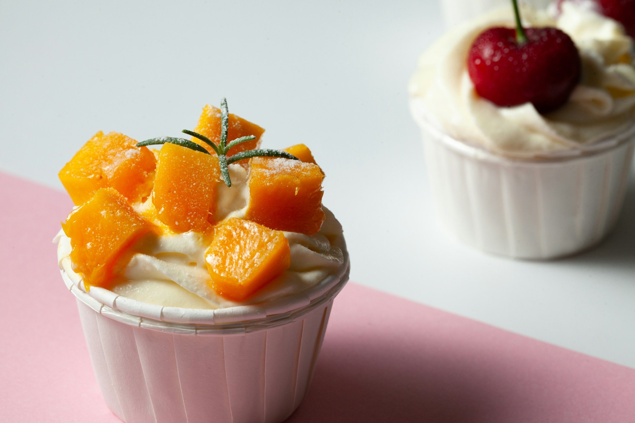Two cups of frozen yogurt with fruit toppings, one with mango pieces and a rosemary sprig in the foreground, and the other with whipped cream and a cherry in the background.