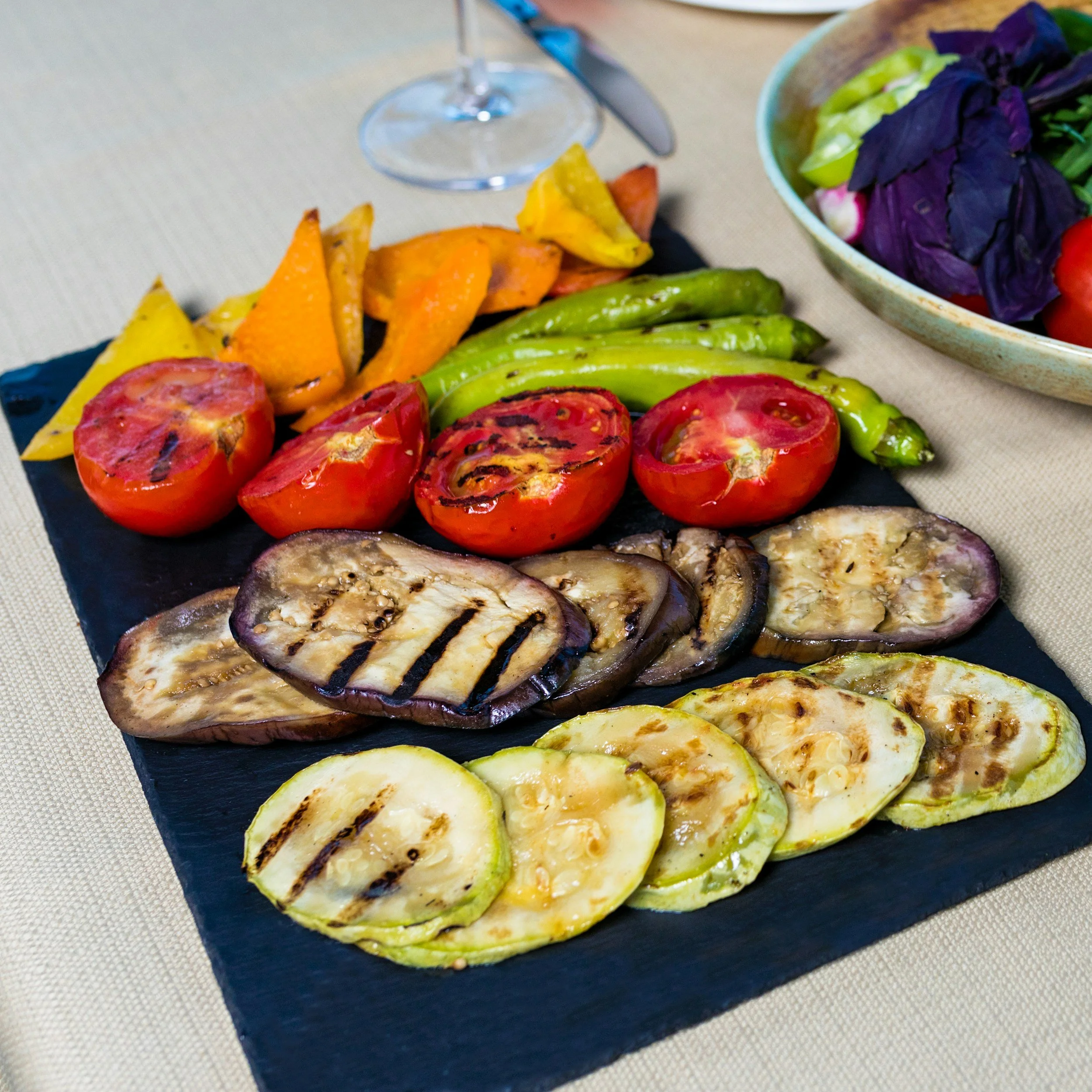 Grilled vegetables including zucchini, eggplant, tomatoes, and green peppers served on a black stone platter at a dining table with a beige tablecloth, a glass of wine, and a bowl of salad in the background.