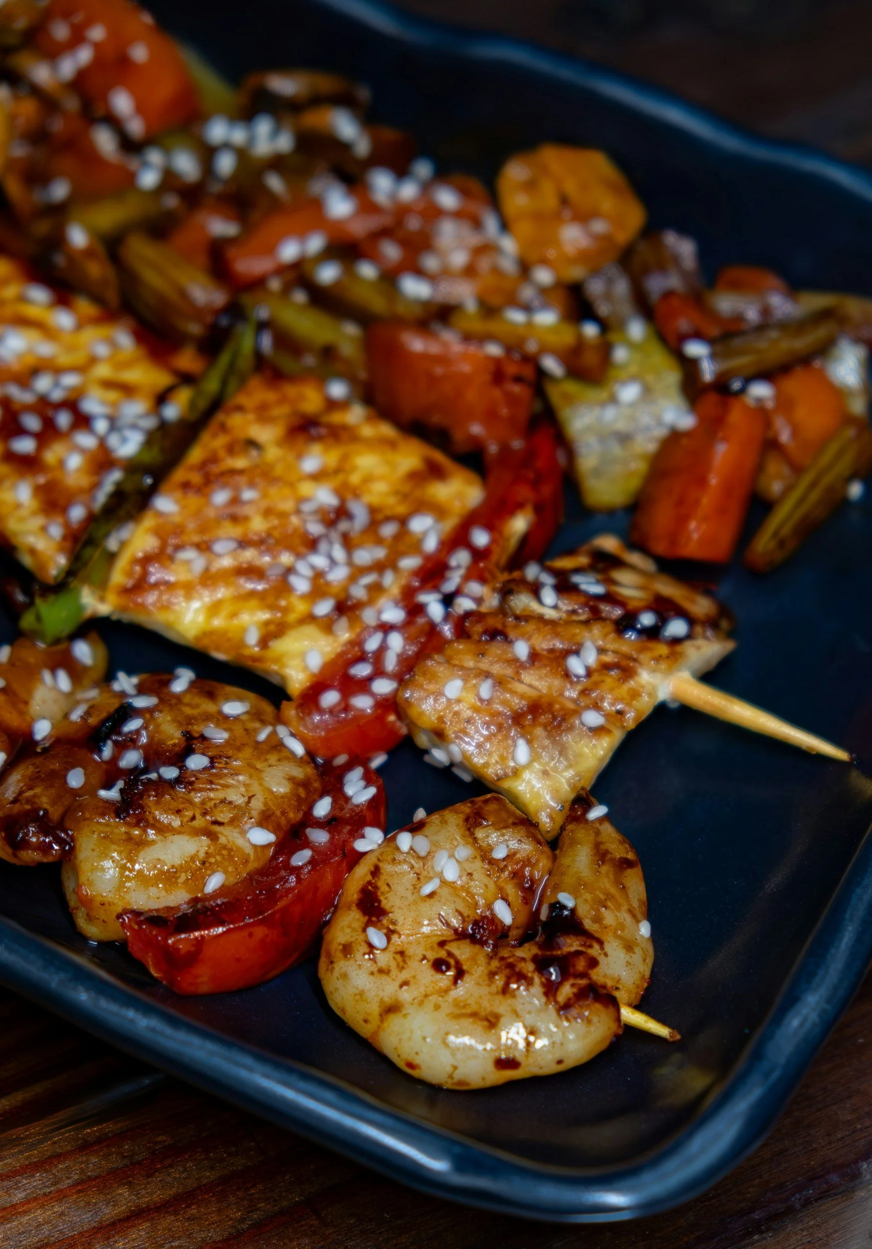 Close-up of grilled chicken skewers with vegetables, topped with sesame seeds on a black plate