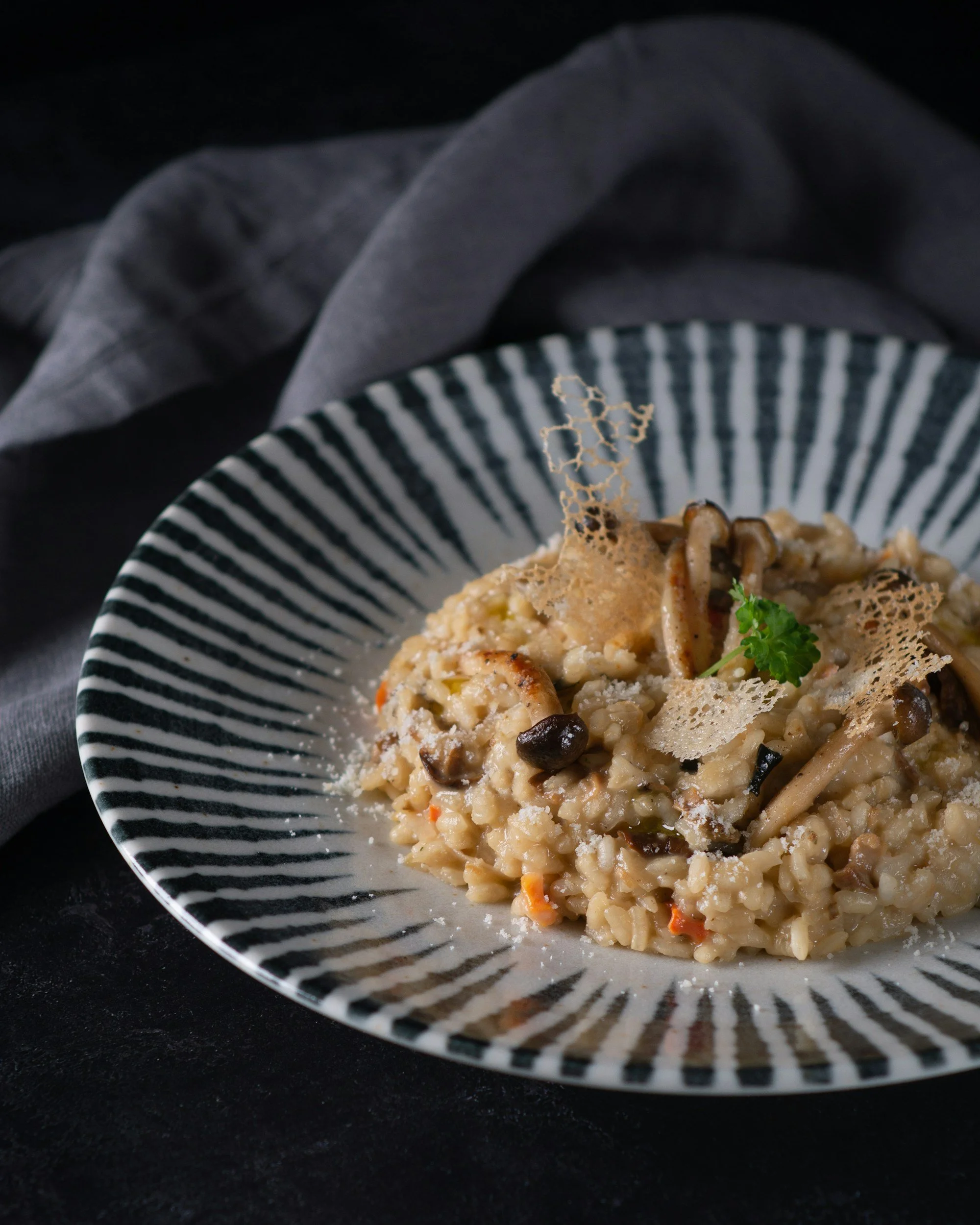 A bowl of mushroom risotto garnished with parmesan cheese, a sprig of parsley, and a lace tuile cookie on a black surface with a gray cloth in the background.