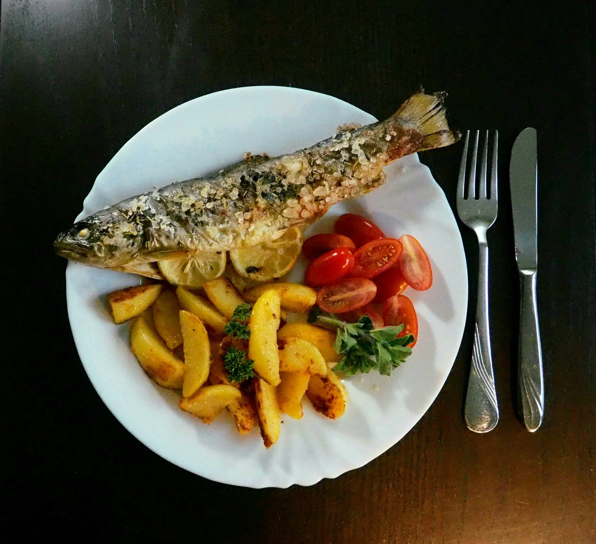 A plate with baked whole fish, roasted potato wedges, cherry tomatoes, and lemon slices, garnished with parsley on a dark table.
