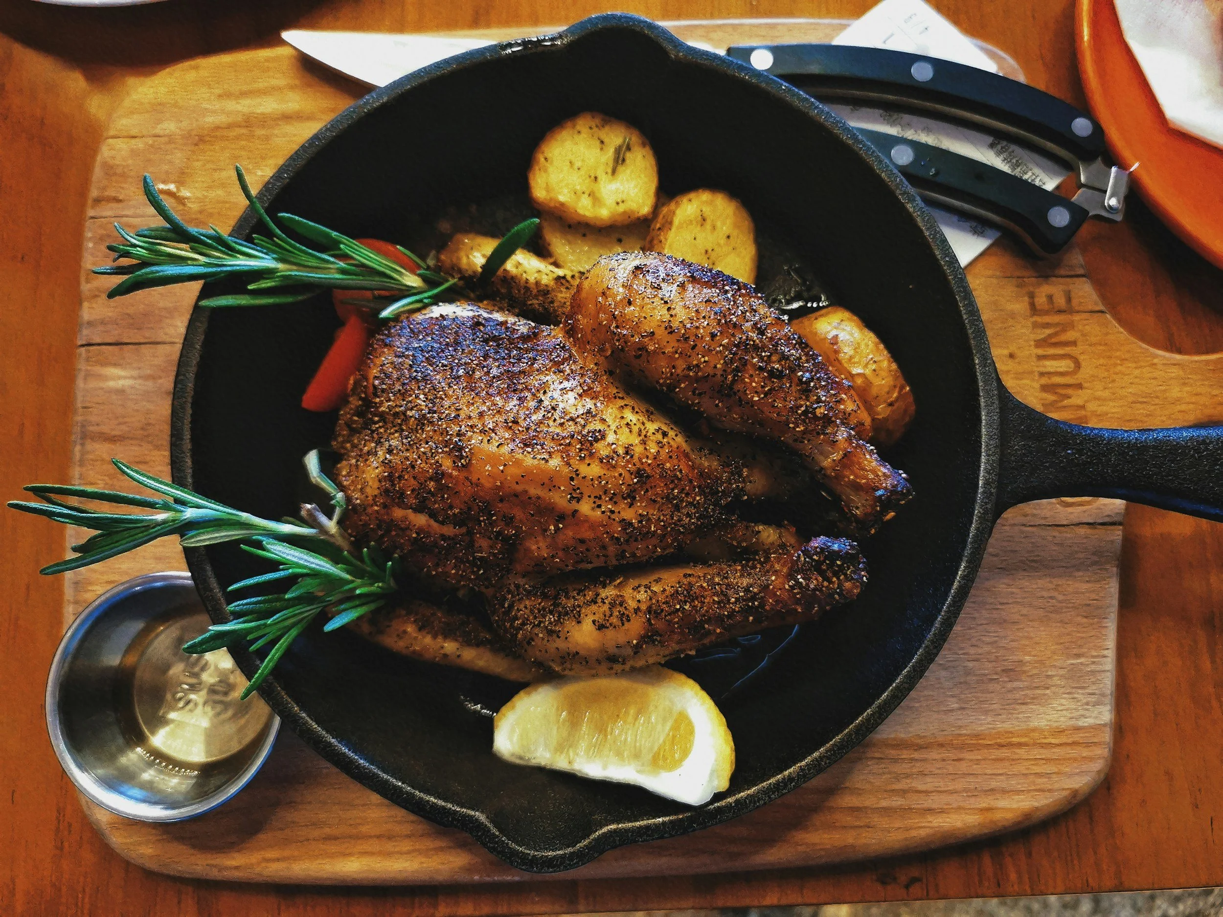 Roast chicken with crispy skin, seasoned with herbs and black pepper, garnished with rosemary sprigs, served with roasted potatoes, lemon wedge, and roasted garlic, in a black cast iron skillet on a wooden serving board.