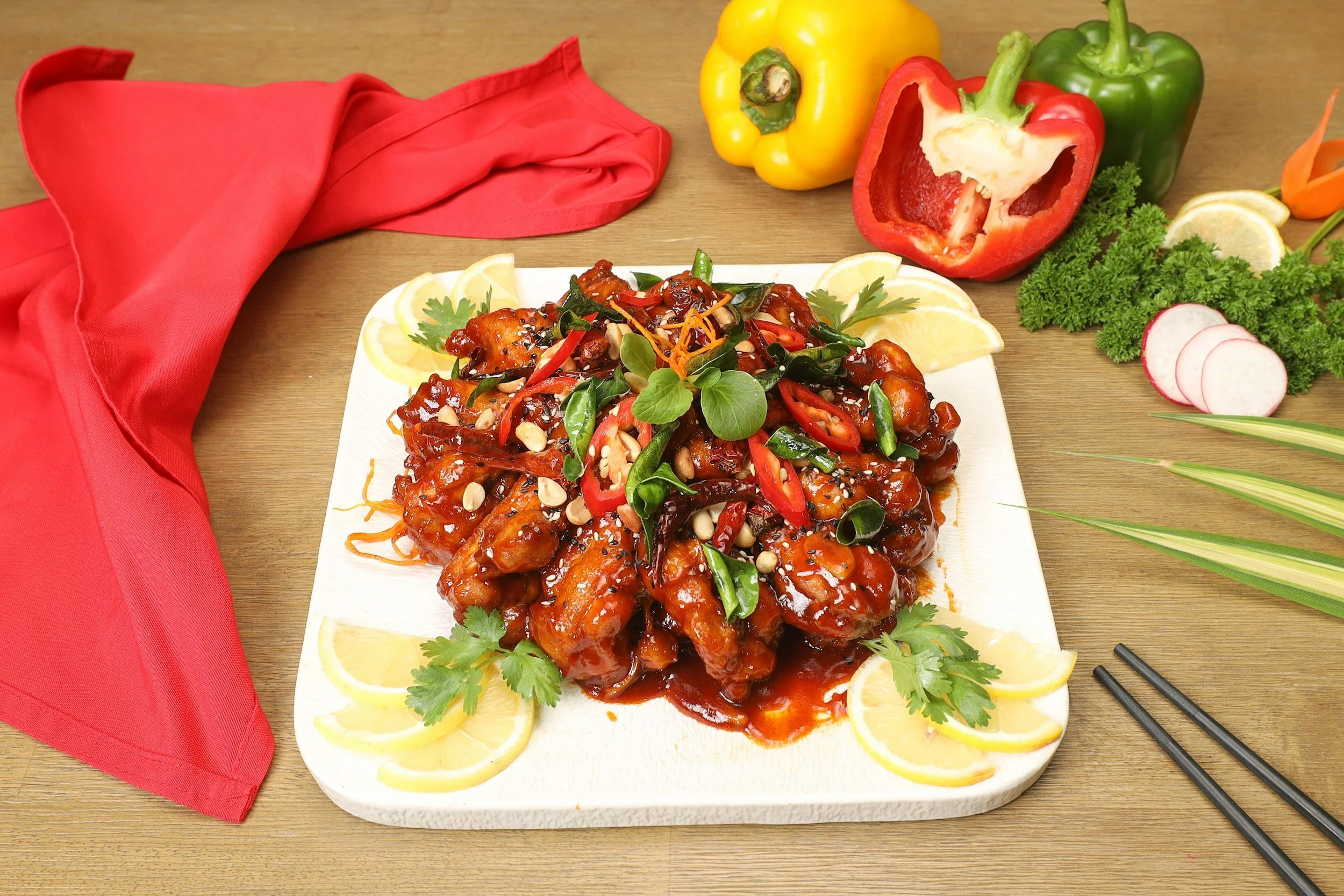 A plate of crispy chicken wings covered in a glossy red sauce, garnished with sliced red chili peppers, green onion, sesame seeds, and fresh herbs, surrounded by lemon wedges, with bell peppers, radish, parsley, and a red cloth napkin on a wooden table.