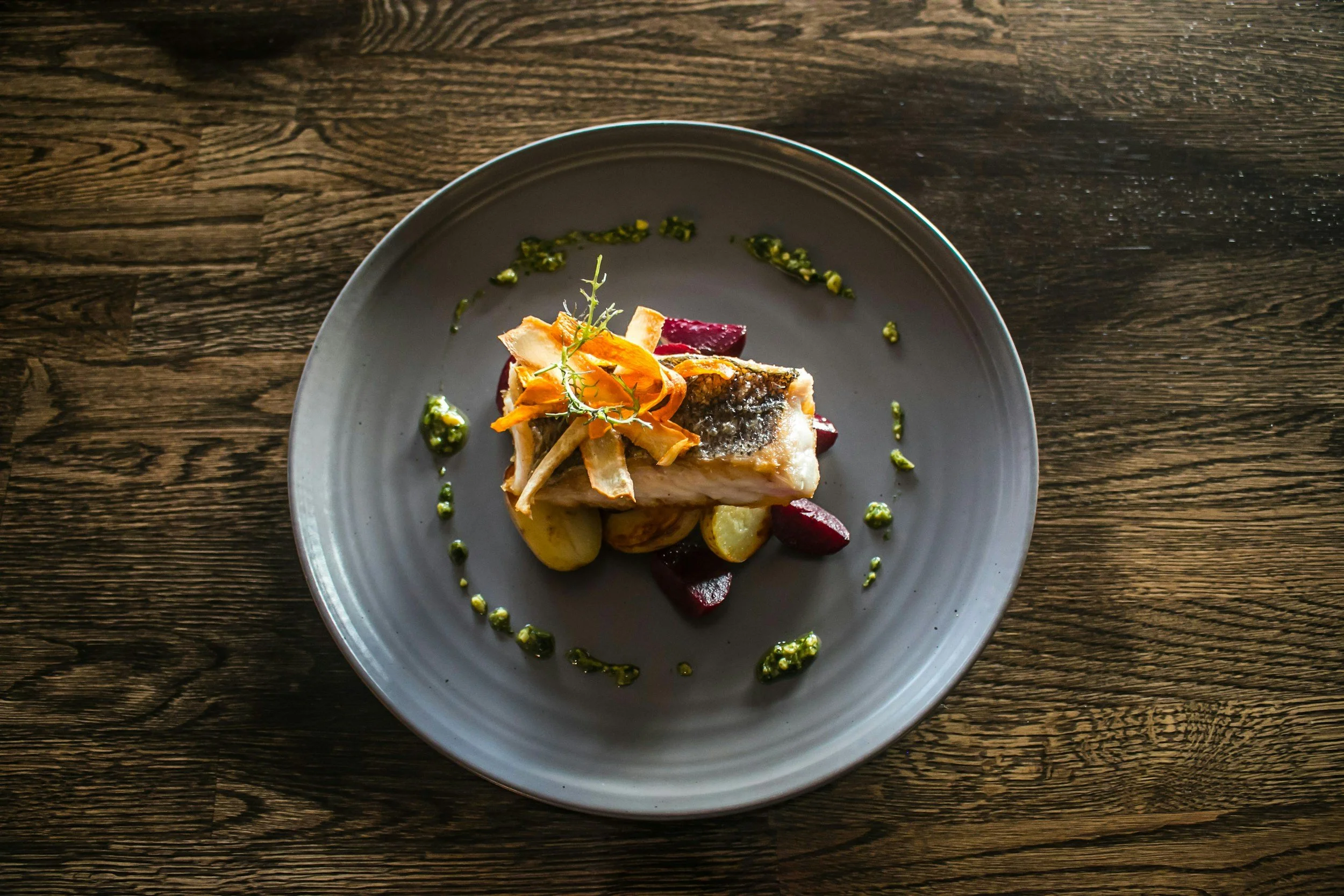 A plated dish with a piece of fish, roasted vegetables, and green sauce on a gray plate, placed on a wooden table.