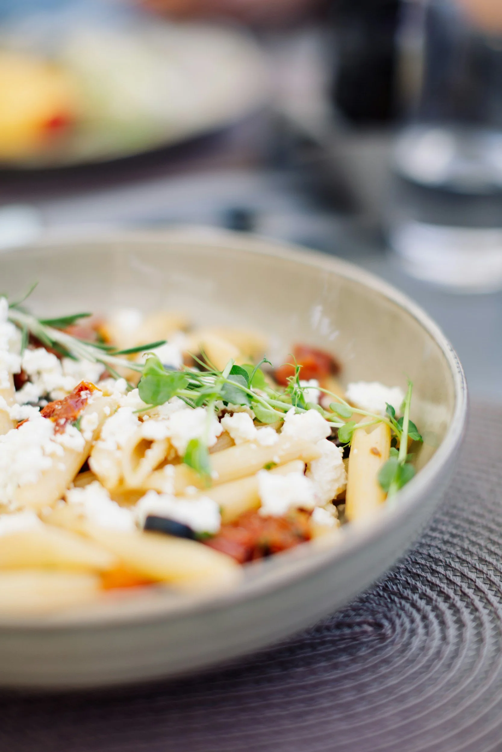 Close-up of a bowl of cooked pasta with crumbled cheese, sun-dried tomatoes, and fresh microgreens on top, placed on a textured surface.