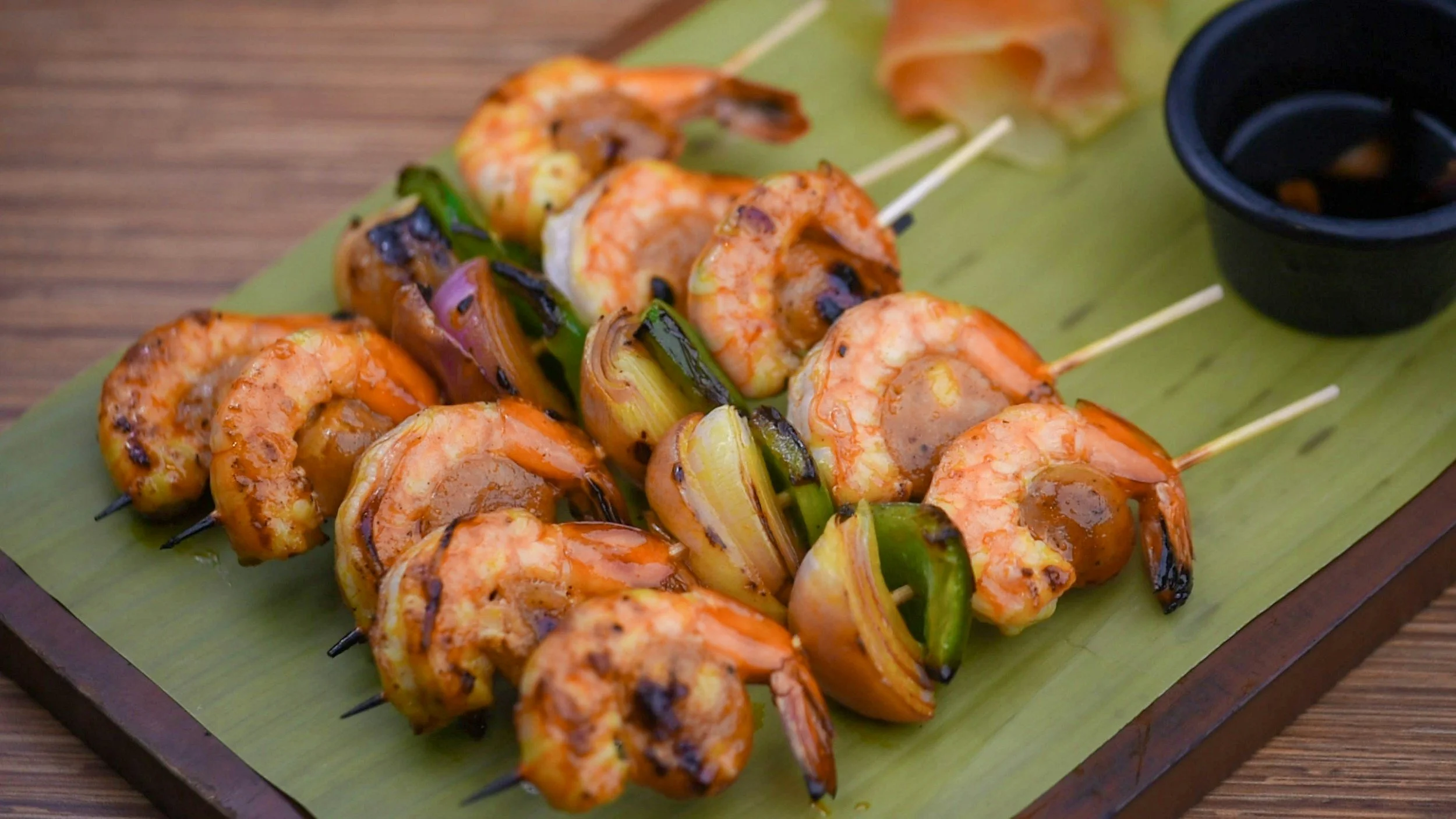 Grilled shrimp and vegetable skewers on a wooden serving board, with a small black dipping bowl of sauce.