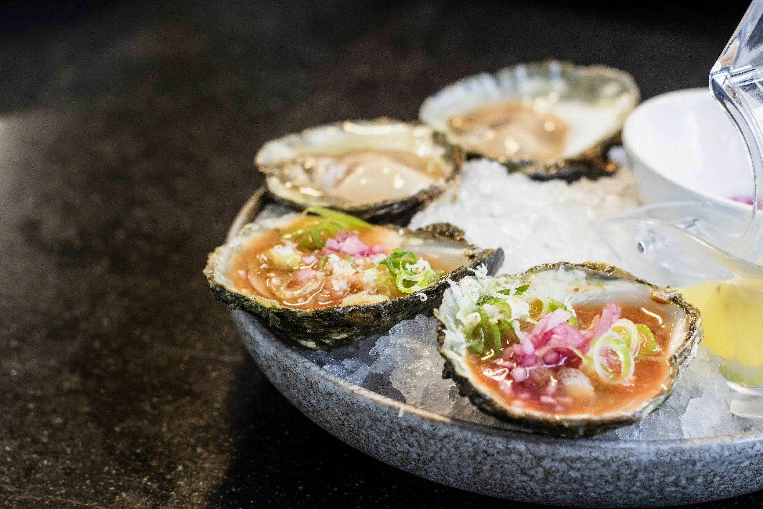 Fresh oysters on a bed of ice with toppings like chopped green onions and pink garnish, served in a bowl.