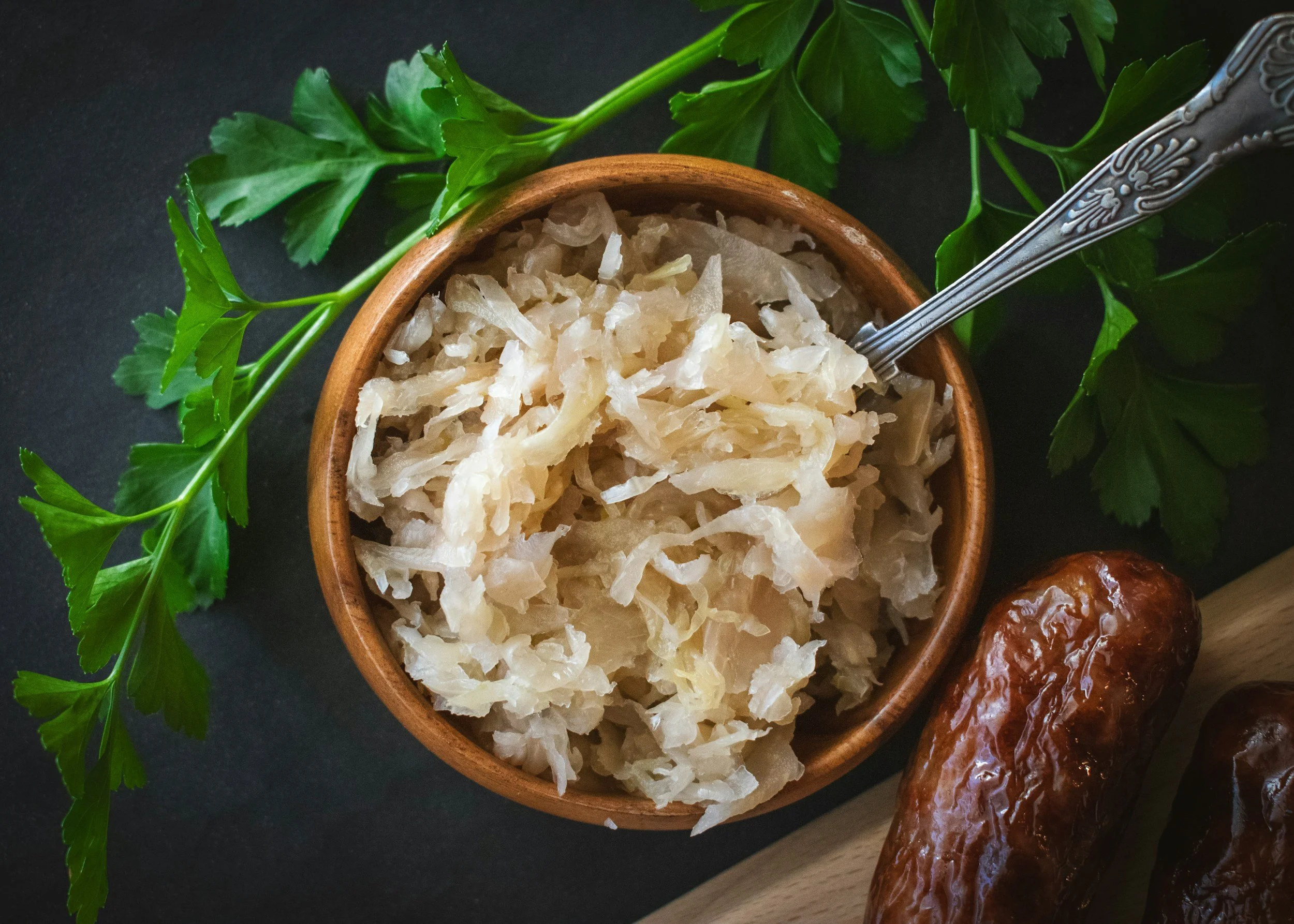 Fermented sauerkraut in a wooden bowl, surrounded by fresh parsley, with dried dates and a silver fork on a black and wood surface.