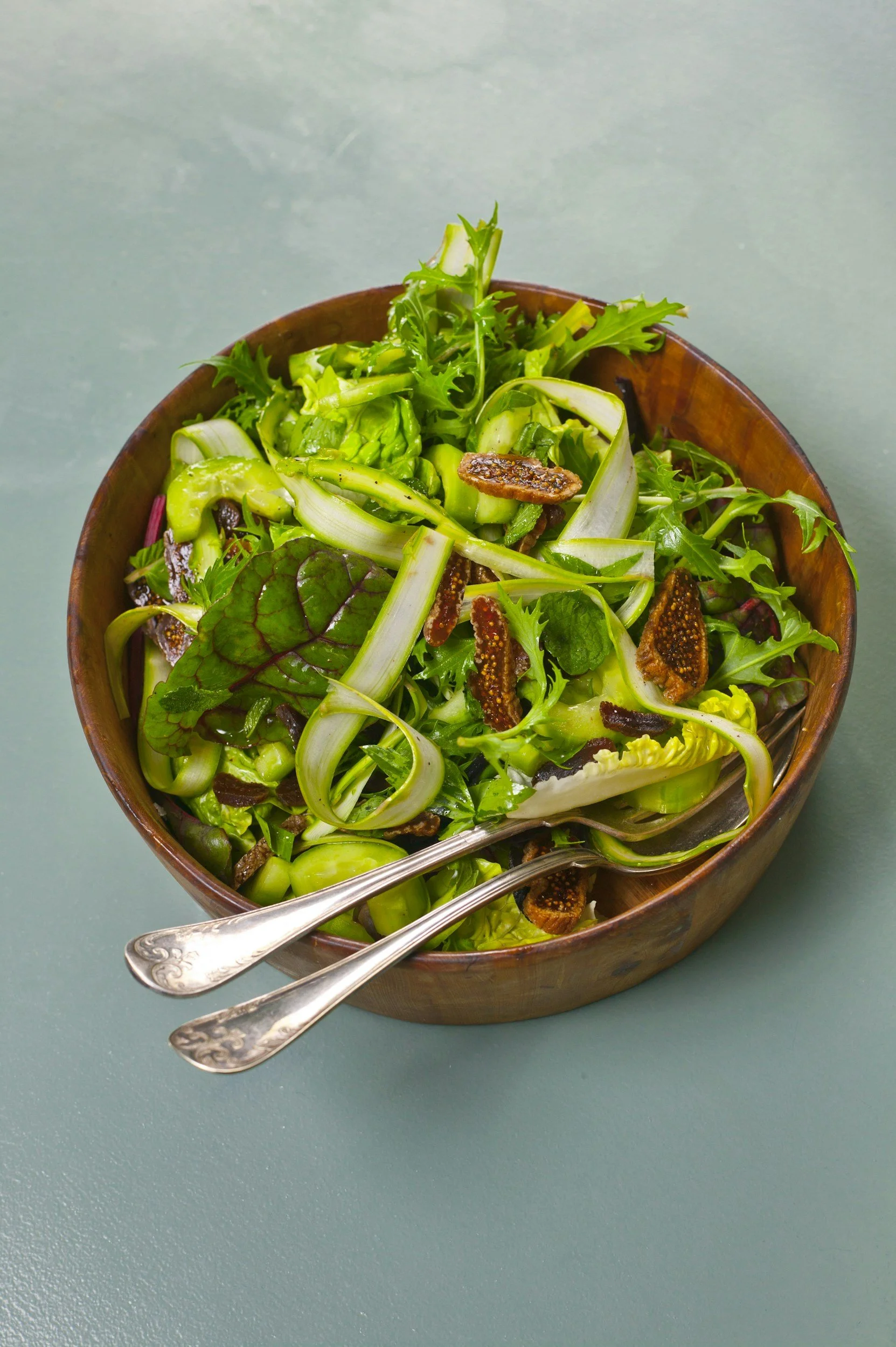 Fresh green salad with mixed greens, sliced vegetables, and figs in a wooden bowl, with a fork and spoon.