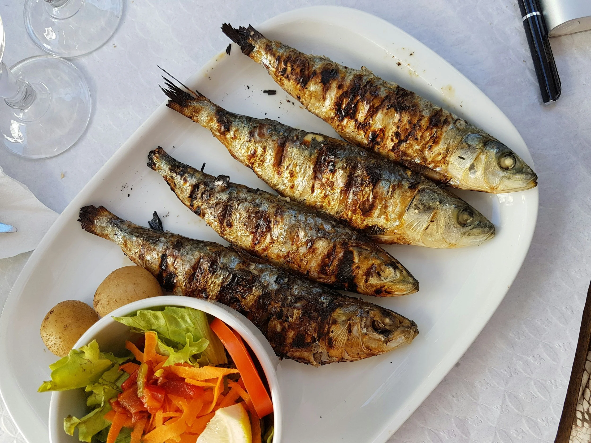Grilled sardines on a white platter with lemon, salad, and boiled potatoes, on a dining table.